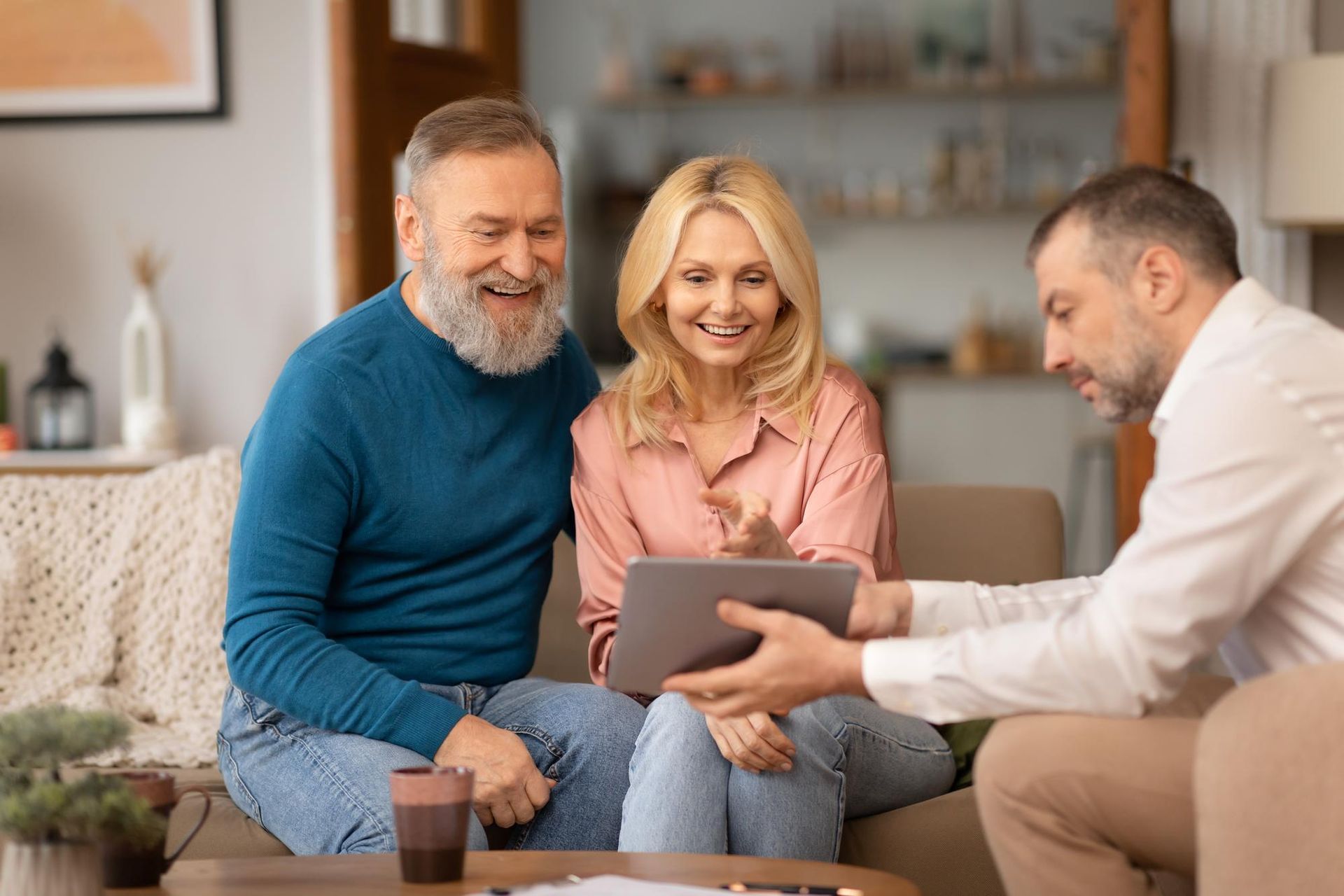 Senior couple using a phone and laptop at a table. Smiling, light-filled room.