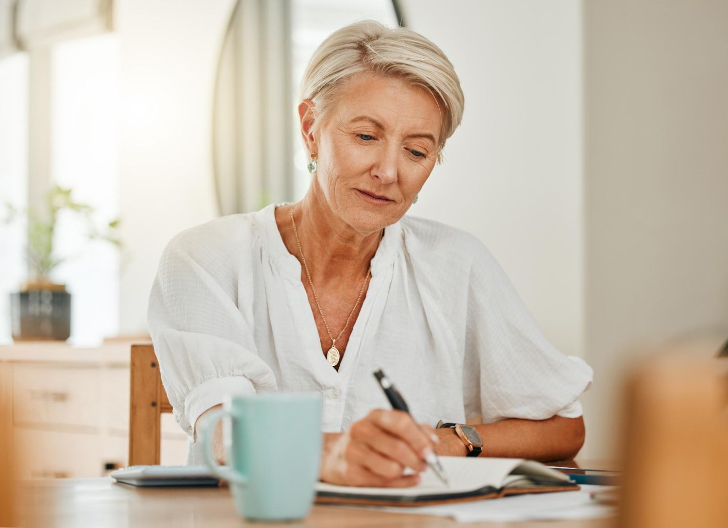 Woman with short gray hair writes in a notebook at a table, with a mug and window in the background.