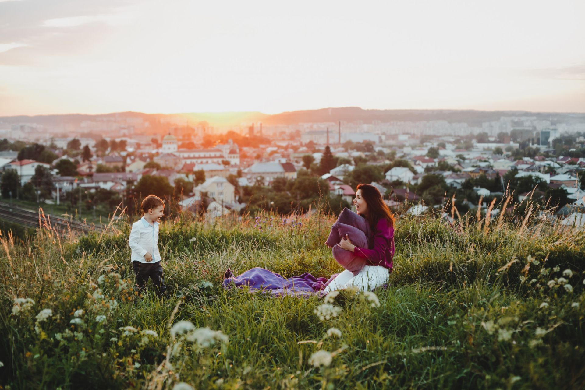 Woman holding baby sits on hill with child in front, overlooking a town at sunset.