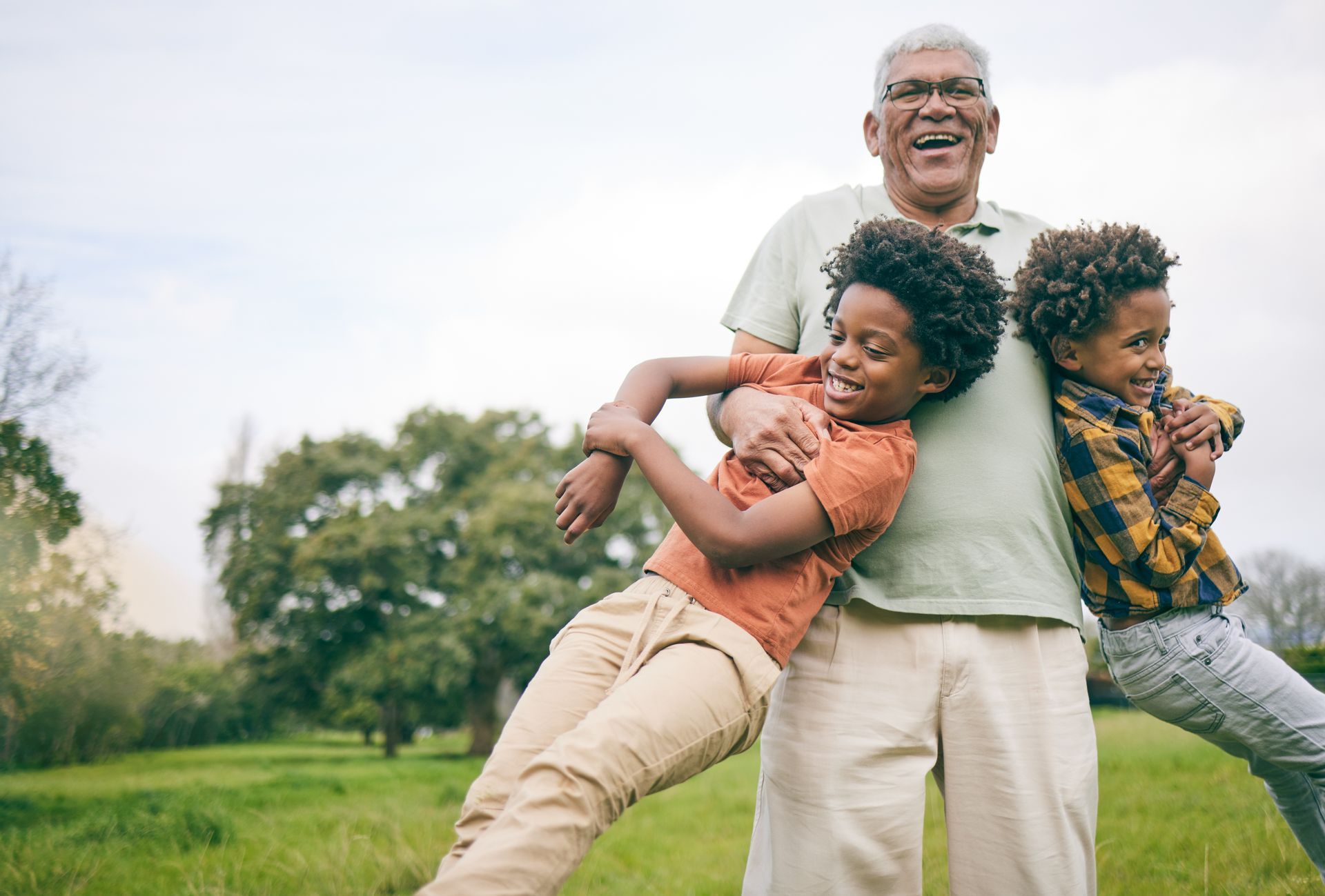 Grandfather smiles as he holds two young boys in a grassy field. The children lean and laugh.