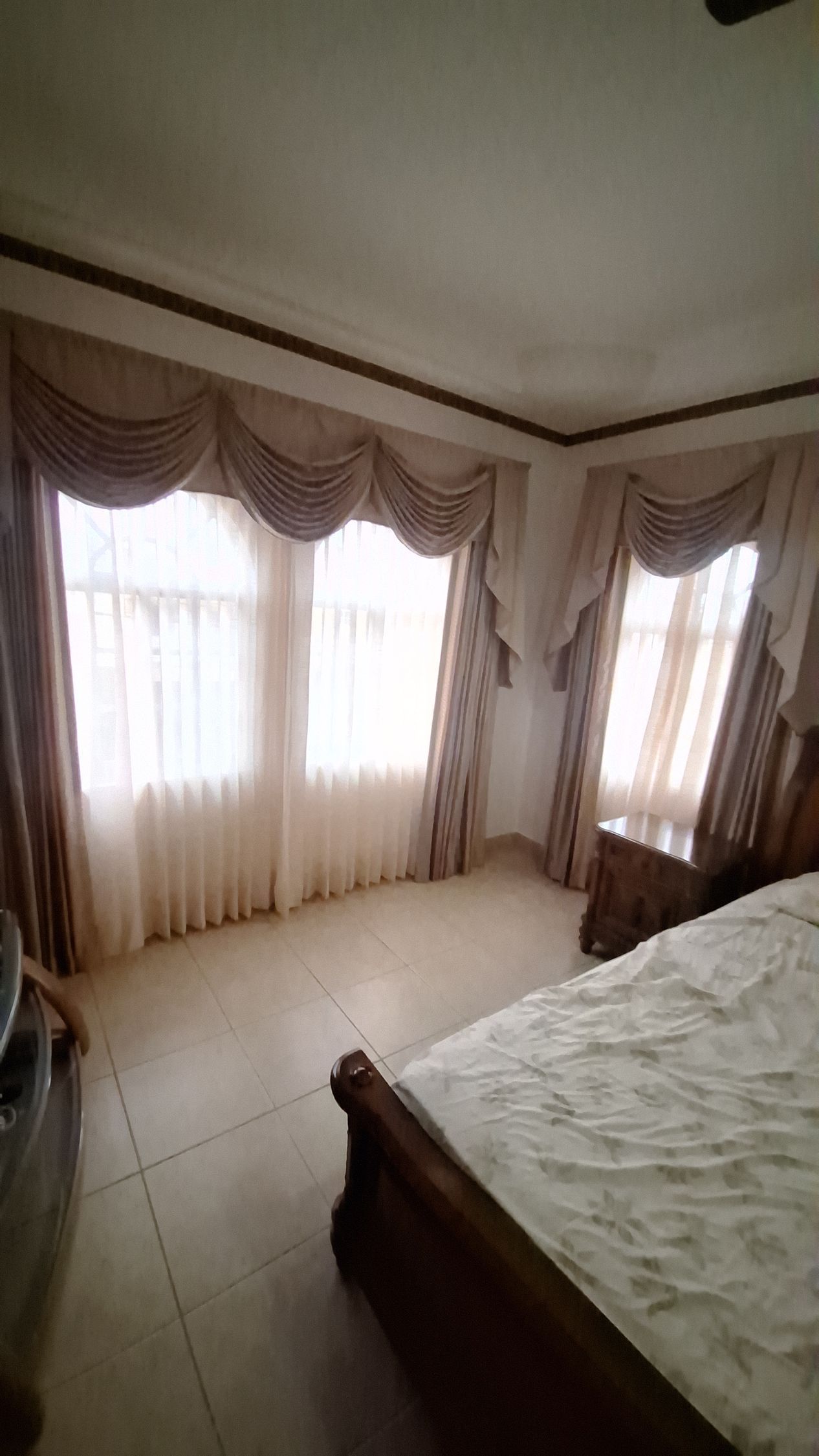 Dormitorio con cortinas claras, cama y mesita de noche. La luz del sol entra a raudales por las ventanas.