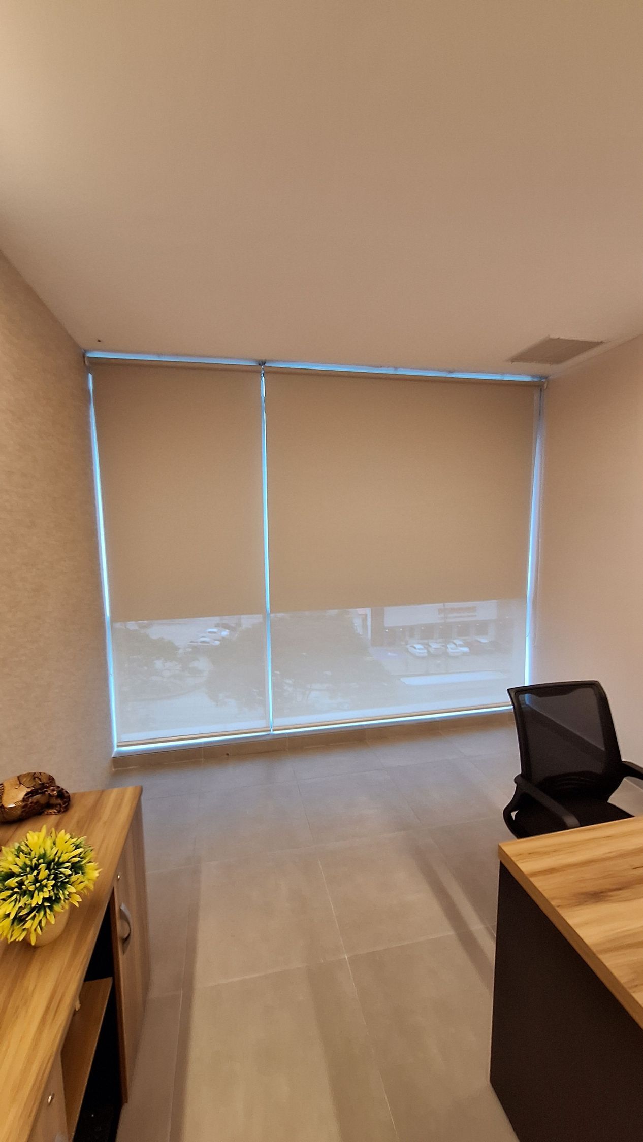 Oficina con persianas cerradas de color marrón, escritorio de madera, silla negra y arreglo floral.