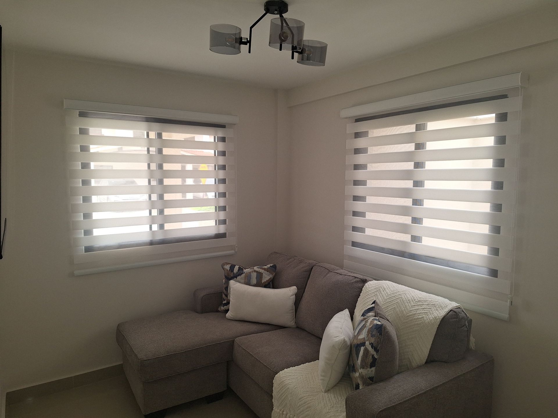 Acogedor rincón de sala de estar con un sofá seccional, ventanas con persianas y una lámpara moderna.