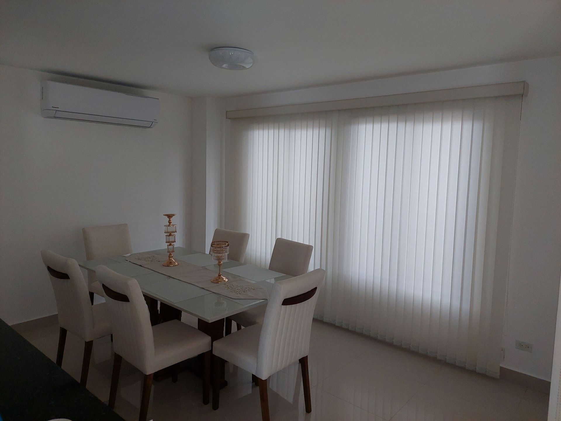 Comedor con mesa de cristal, seis sillas, persianas verticales y aire acondicionado.
