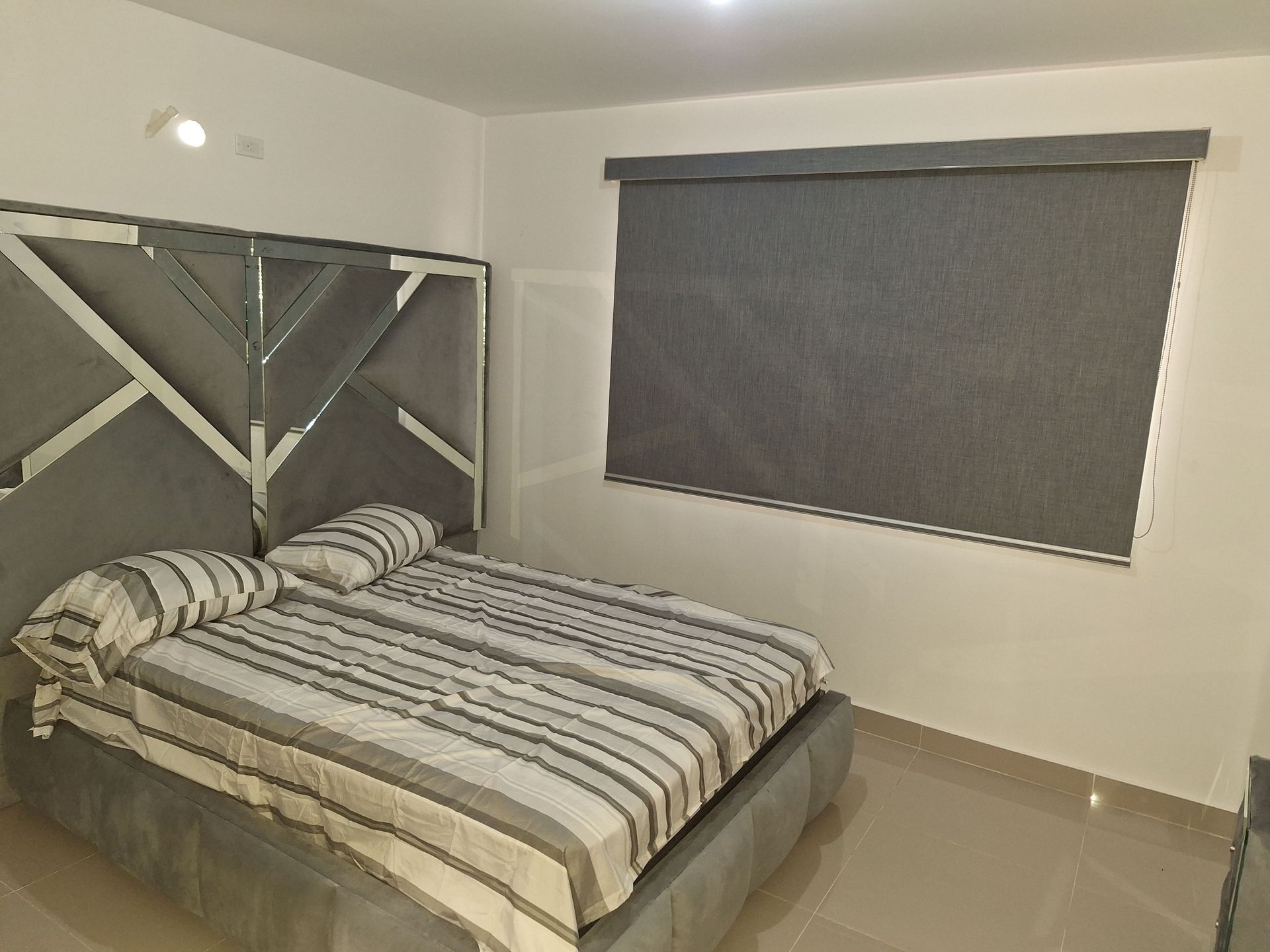 Dormitorio con cama, cabecero gris y ventana con persiana gris cerrada.