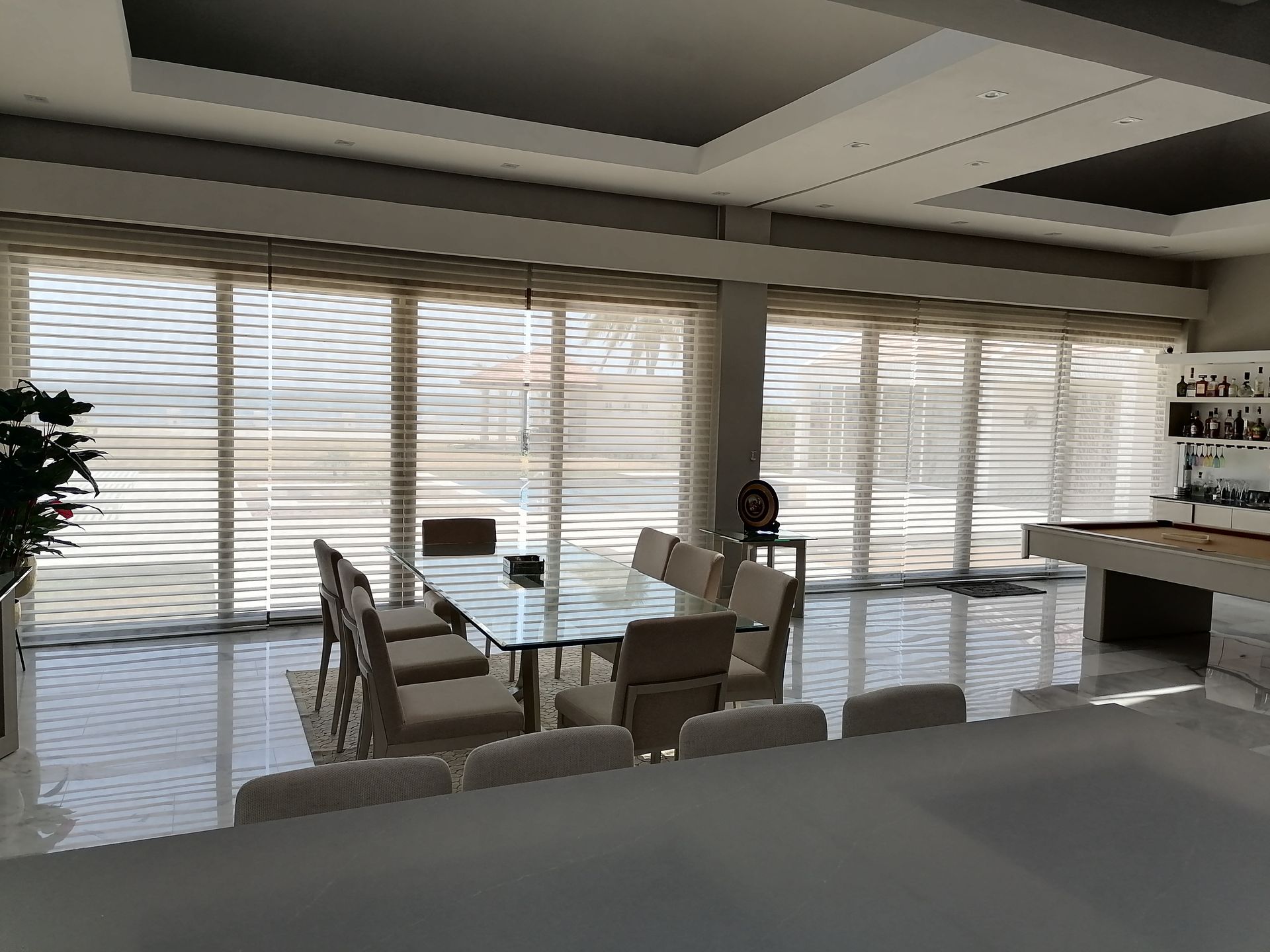 Comedor con mesa de cristal, sillas color beige, amplios ventanales con persianas y barra.