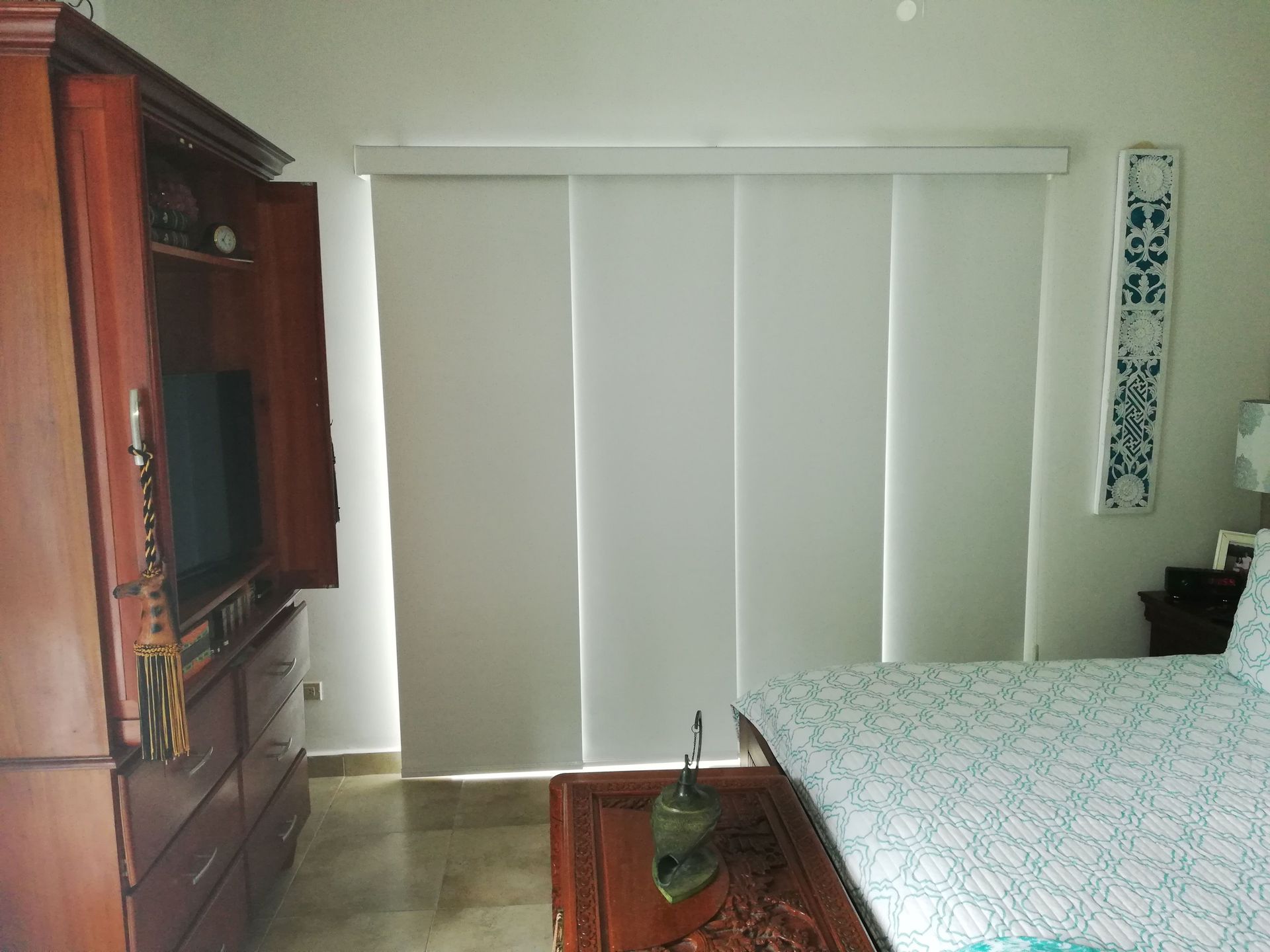 Dormitorio con persianas cerradas, cómoda de madera y cama con ropa de cama estampada.