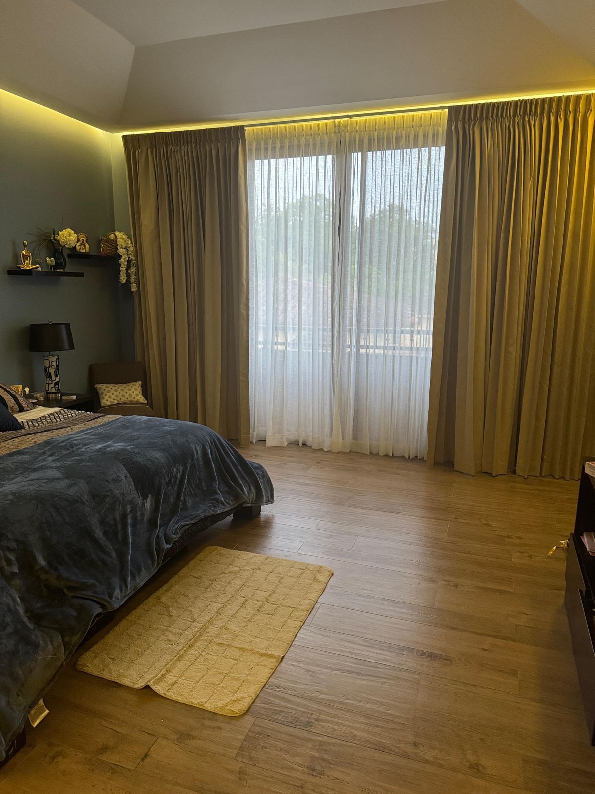Dormitorio con cortinas color canela, cortinas blancas transparentes, piso de madera y una cama con edredón azul.