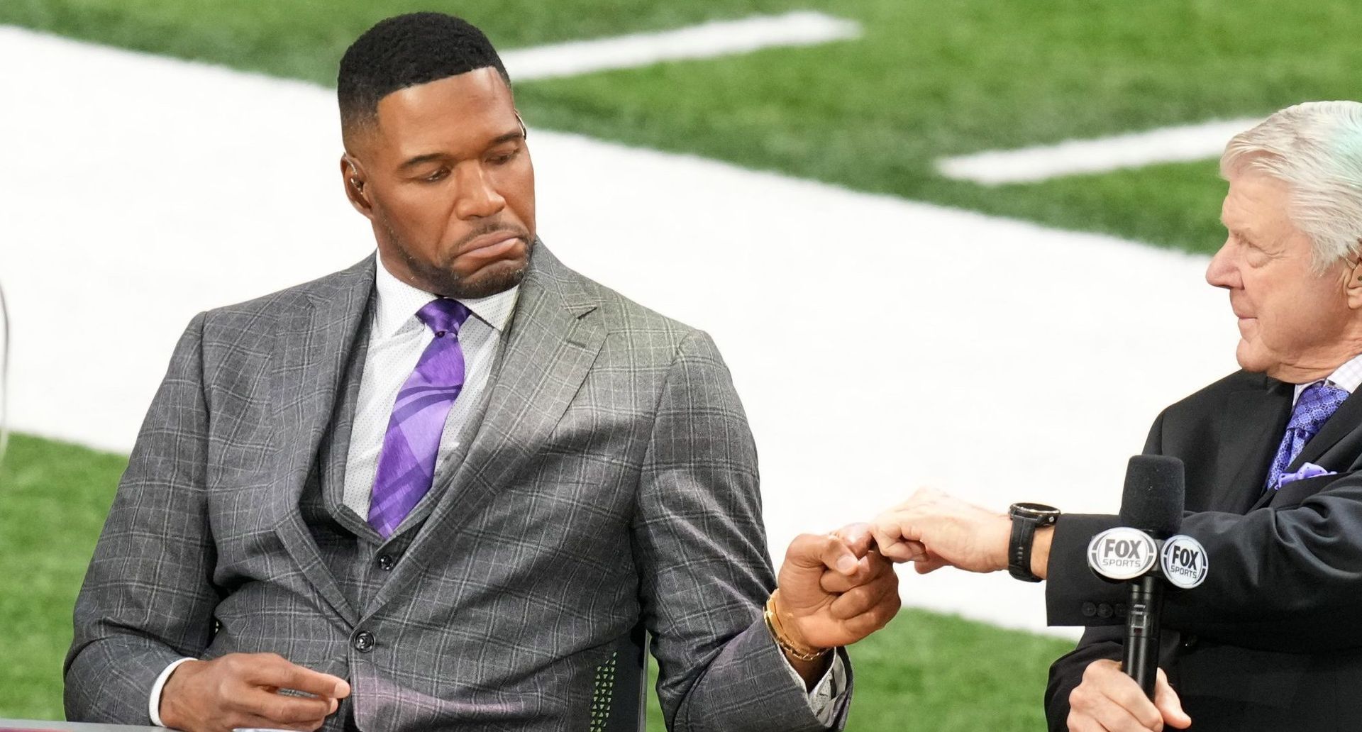 A man in a suit and tie is sitting next to another man on a football field.