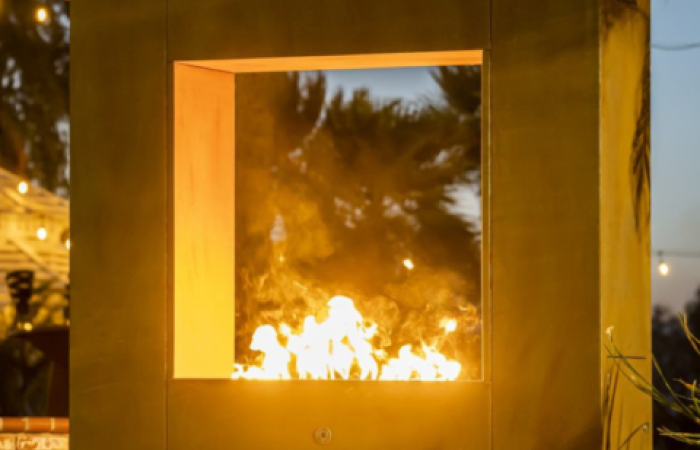 A fireplace is lit up in a square window with trees in the background.