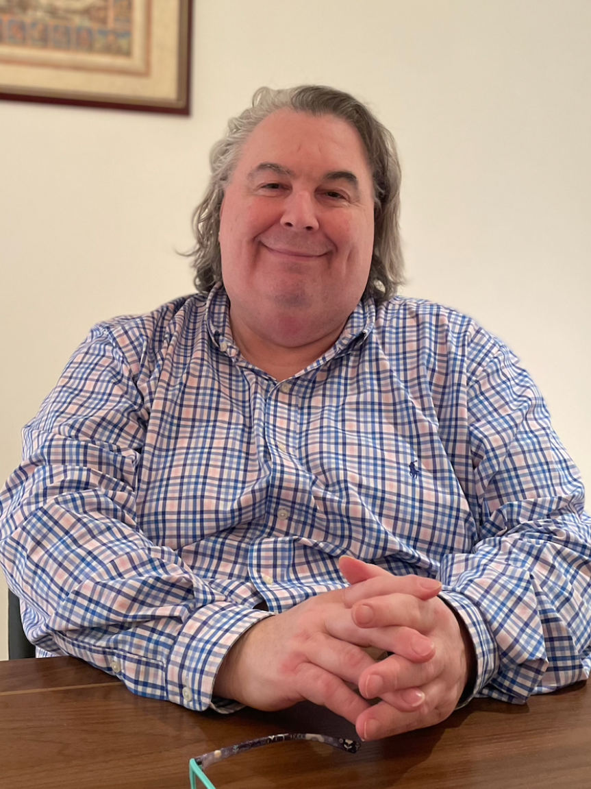 A man with gray hair and a blue-and-white checked shirt sits at a wooden table, smiling. Glasses lie on the table.
