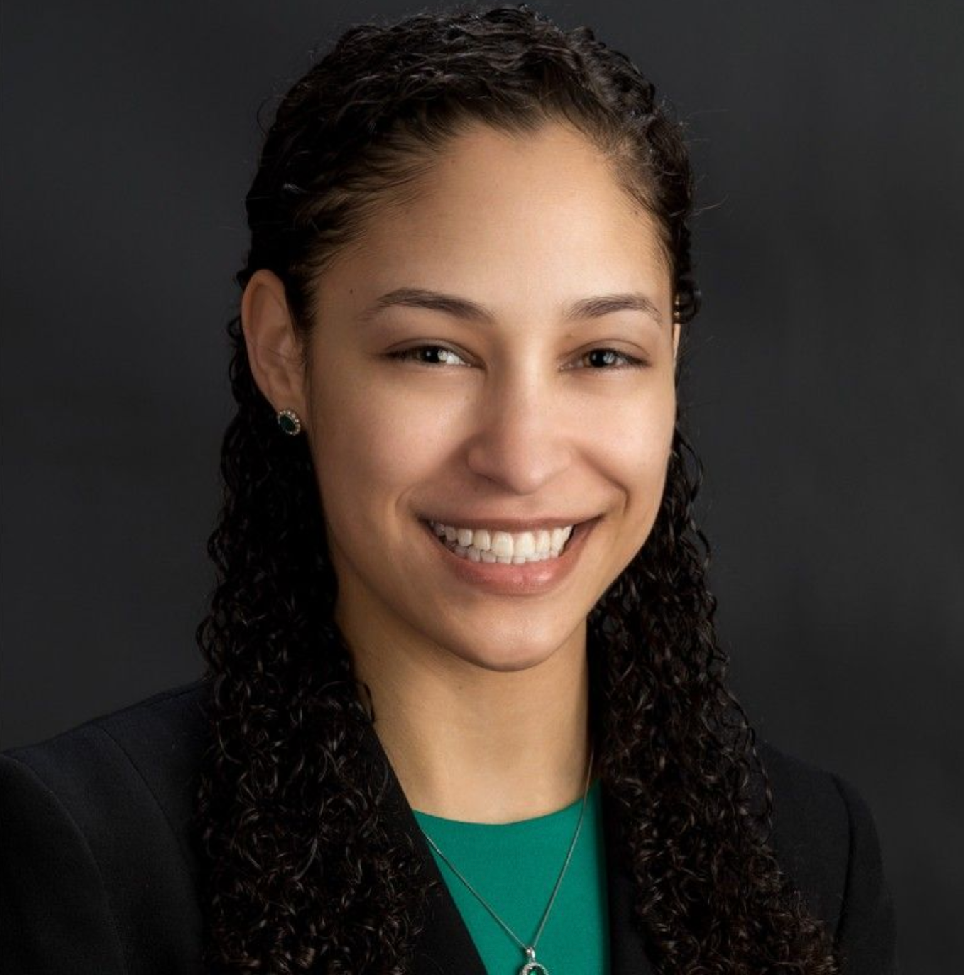 A woman with curly hair is smiling and wearing a green shirt and a black jacket.