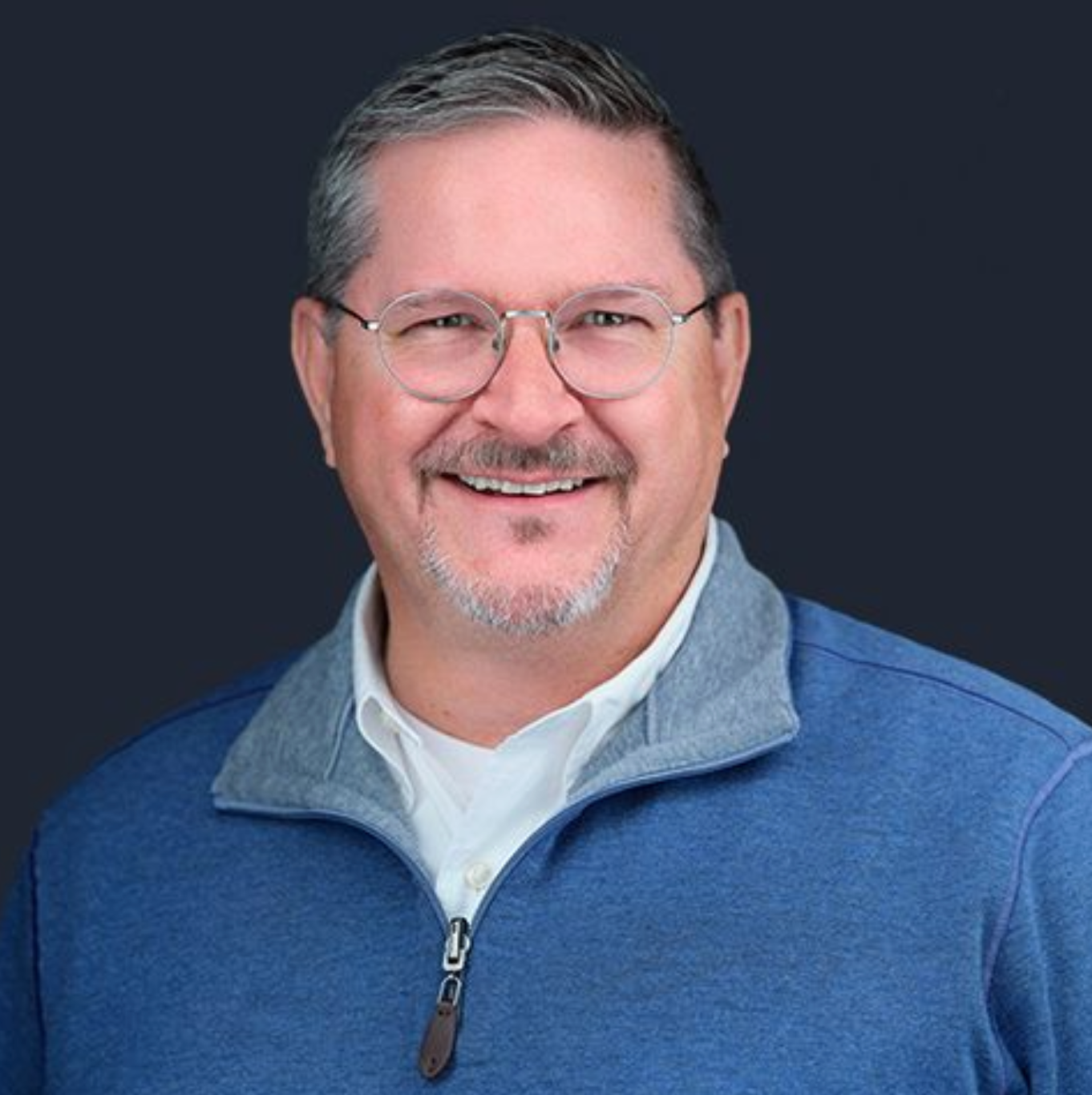 A man wearing glasses and a blue sweater is smiling for the camera.
