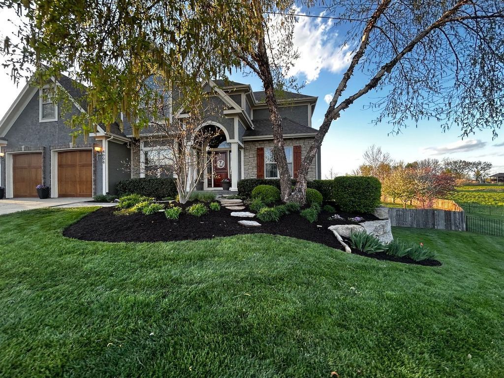 House with green lawn, trees, and dark mulch around bushes, a sunny sky.