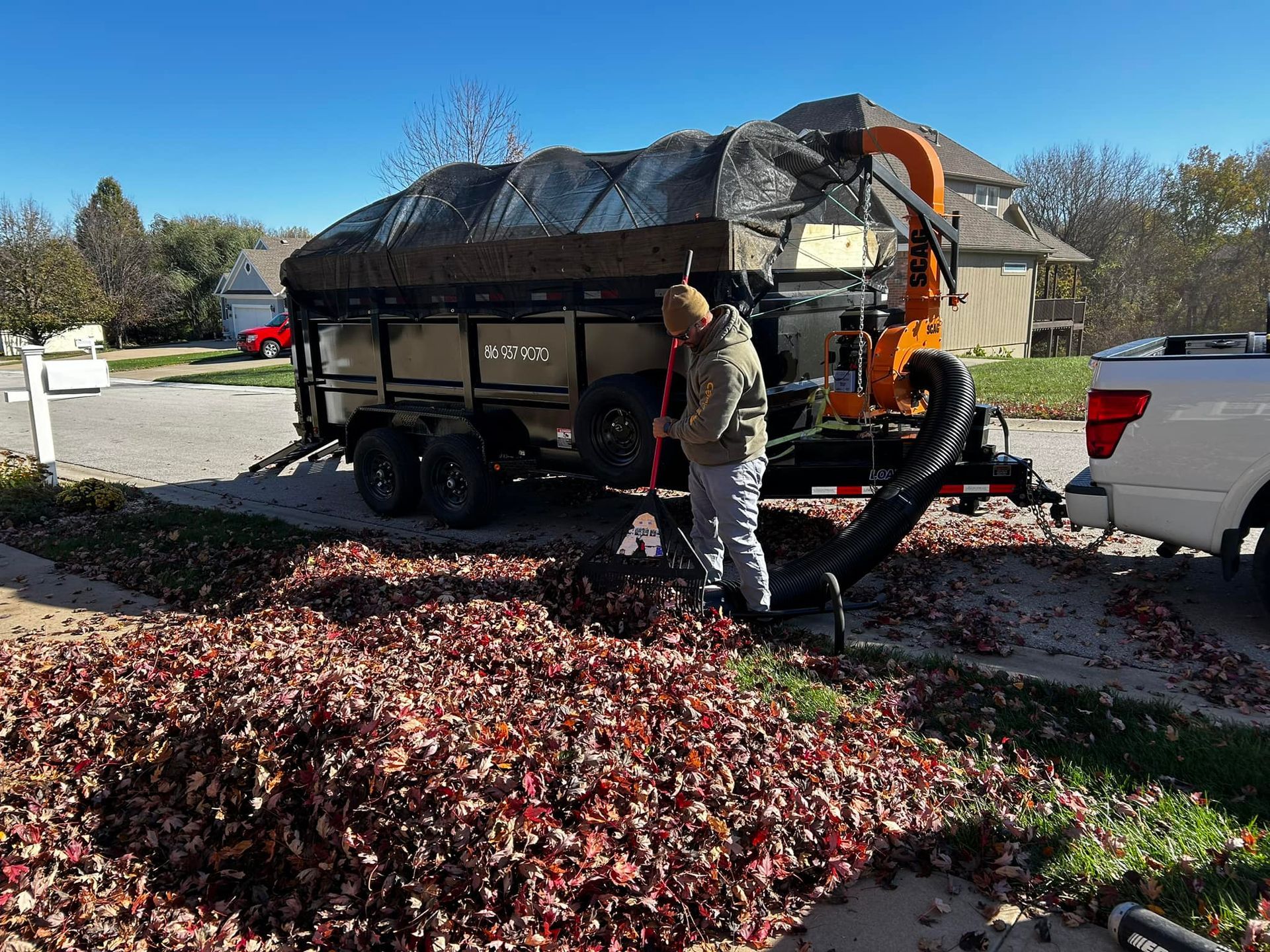 Man rakes leaves into a trailer with a vacuum attached. Leaves are piled on a driveway.