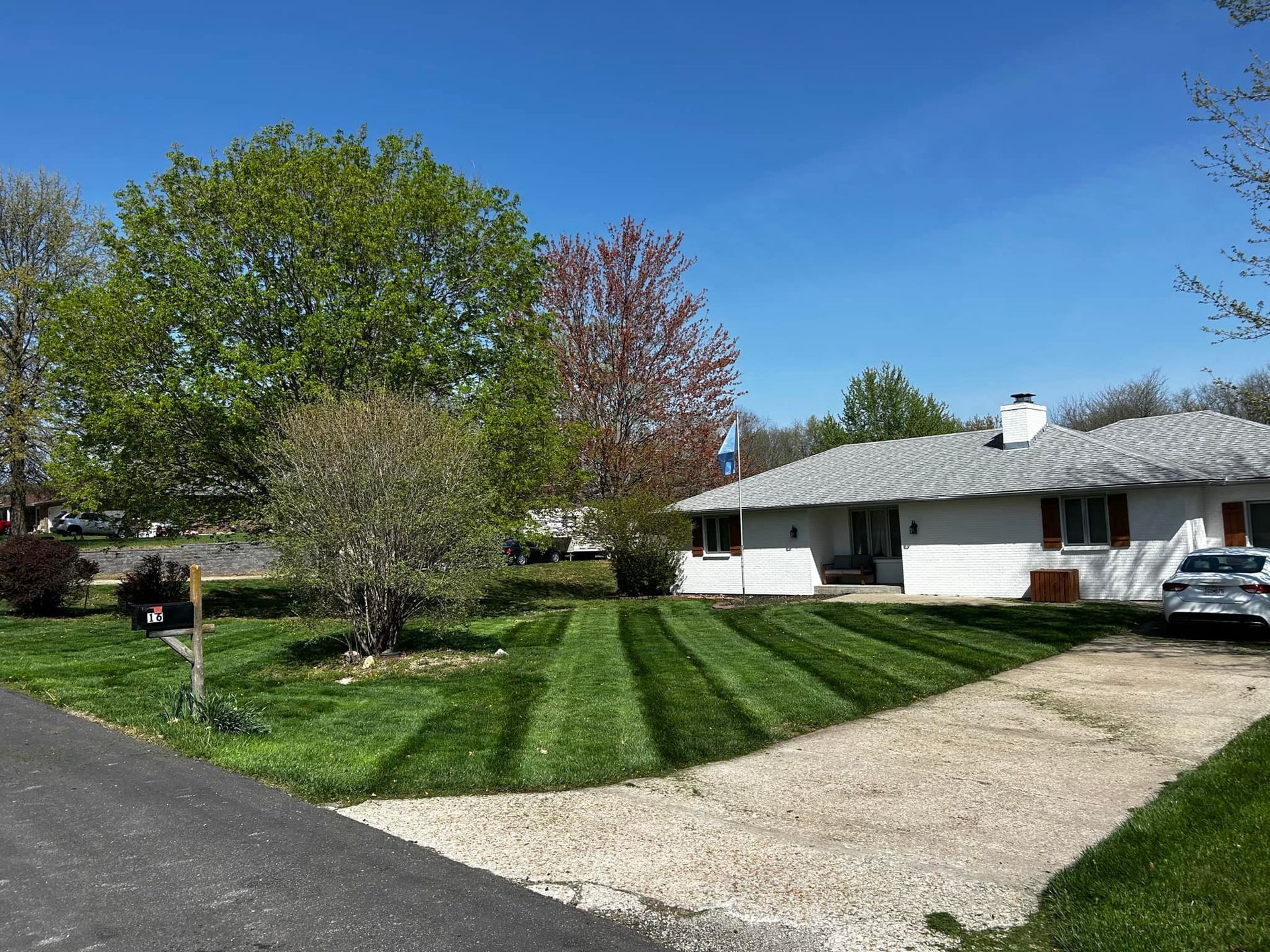 White house with striped green lawn, trees, and a driveway on a sunny day.