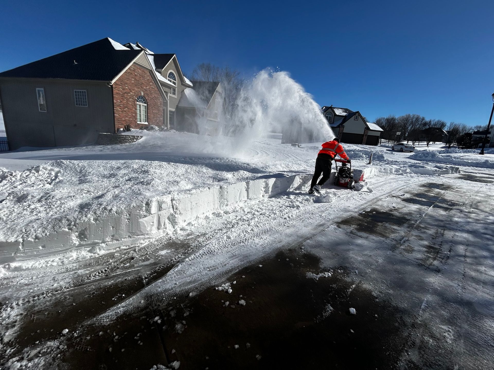 Person using a snow blower, clearing snow from a driveway in front of a house on a sunny day.
