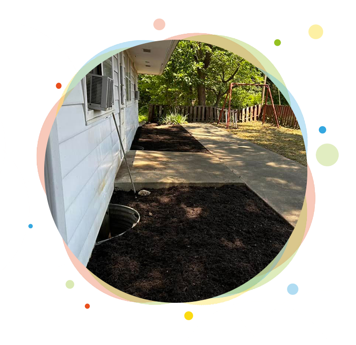 A house exterior with newly mulched garden beds along a concrete walkway, trees in background.