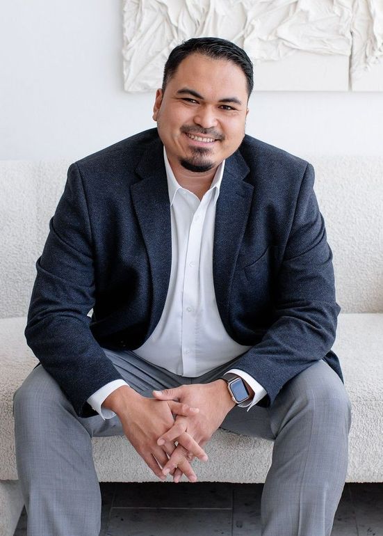 A male realtor wearing a blue suit is sitting on a sofa. The background is a wall that is painted white with art hanging on it.