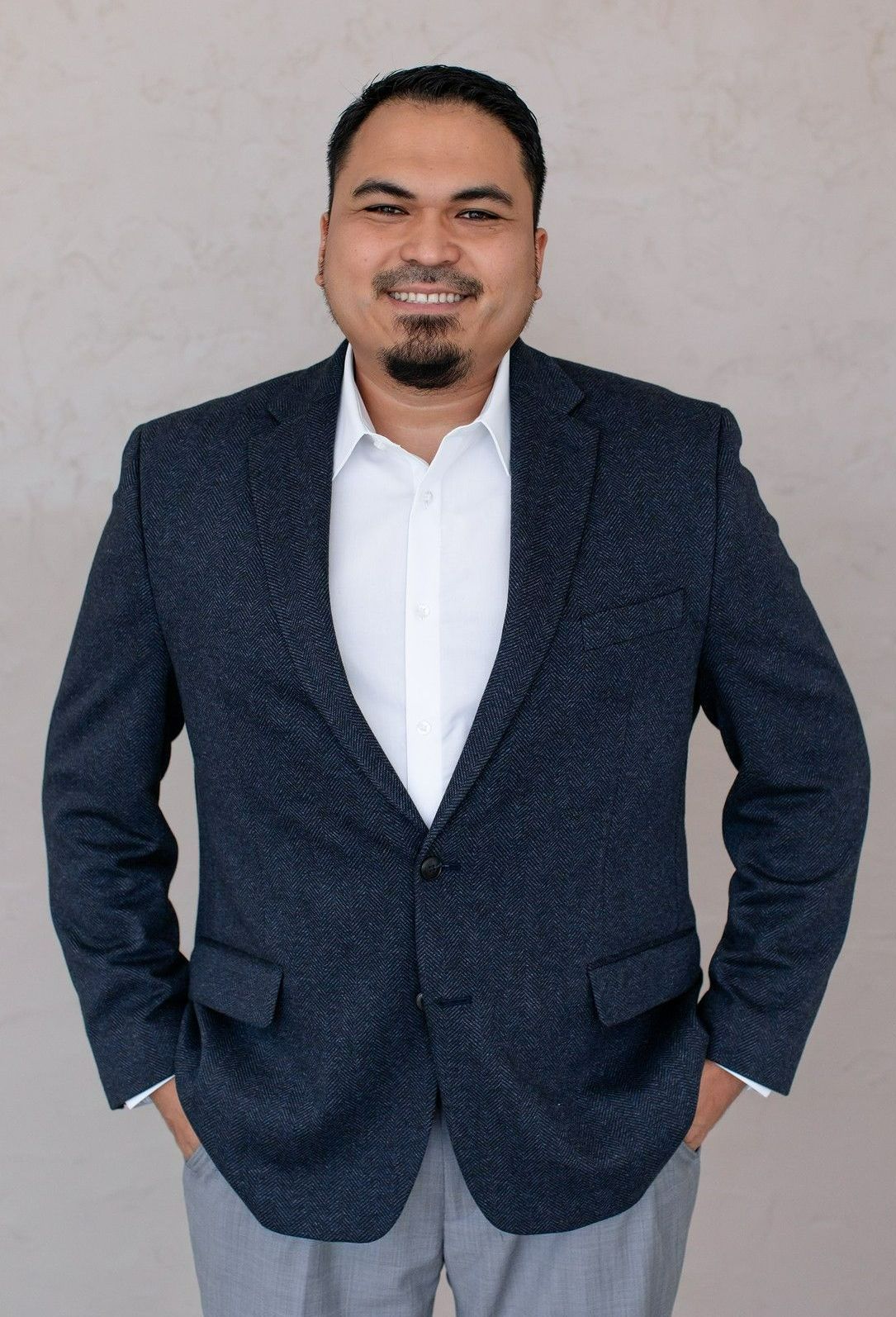 A male realtor wearing a blue suit is standing on a gray wall.