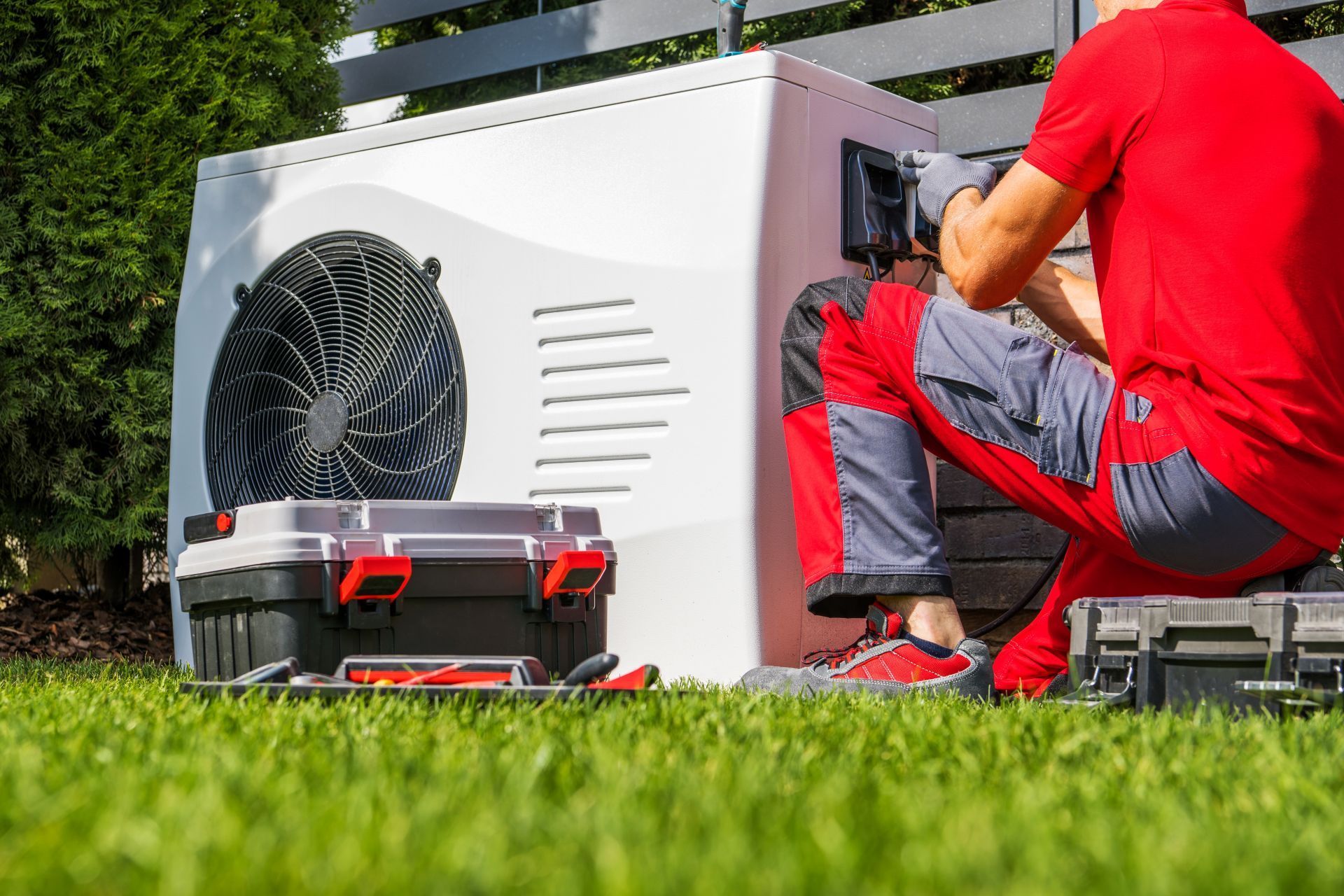 Person in red shirt and gray pants works on an outdoor HVAC unit, tools in a toolbox on the grass.