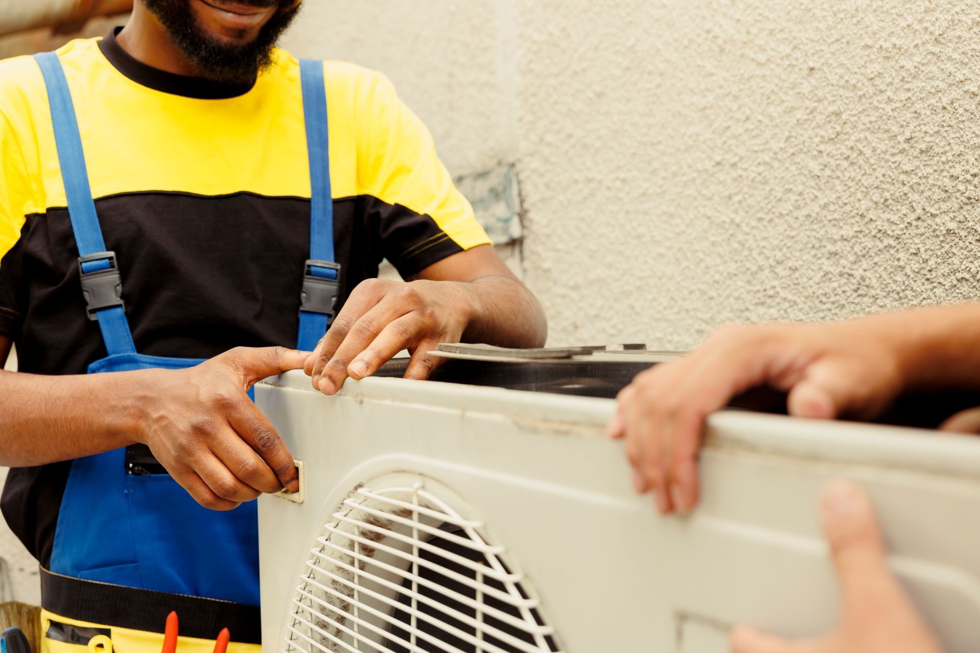 Two technicians installing an air conditioning unit outside a building. One wears blue overalls and yellow shirt.