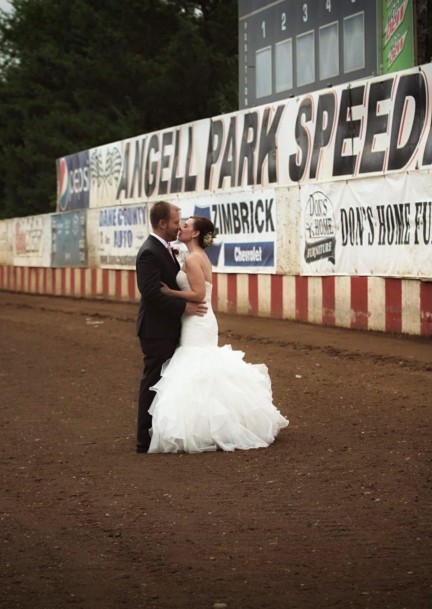A bride and groom kissing in front of a sign that says angell park speedway