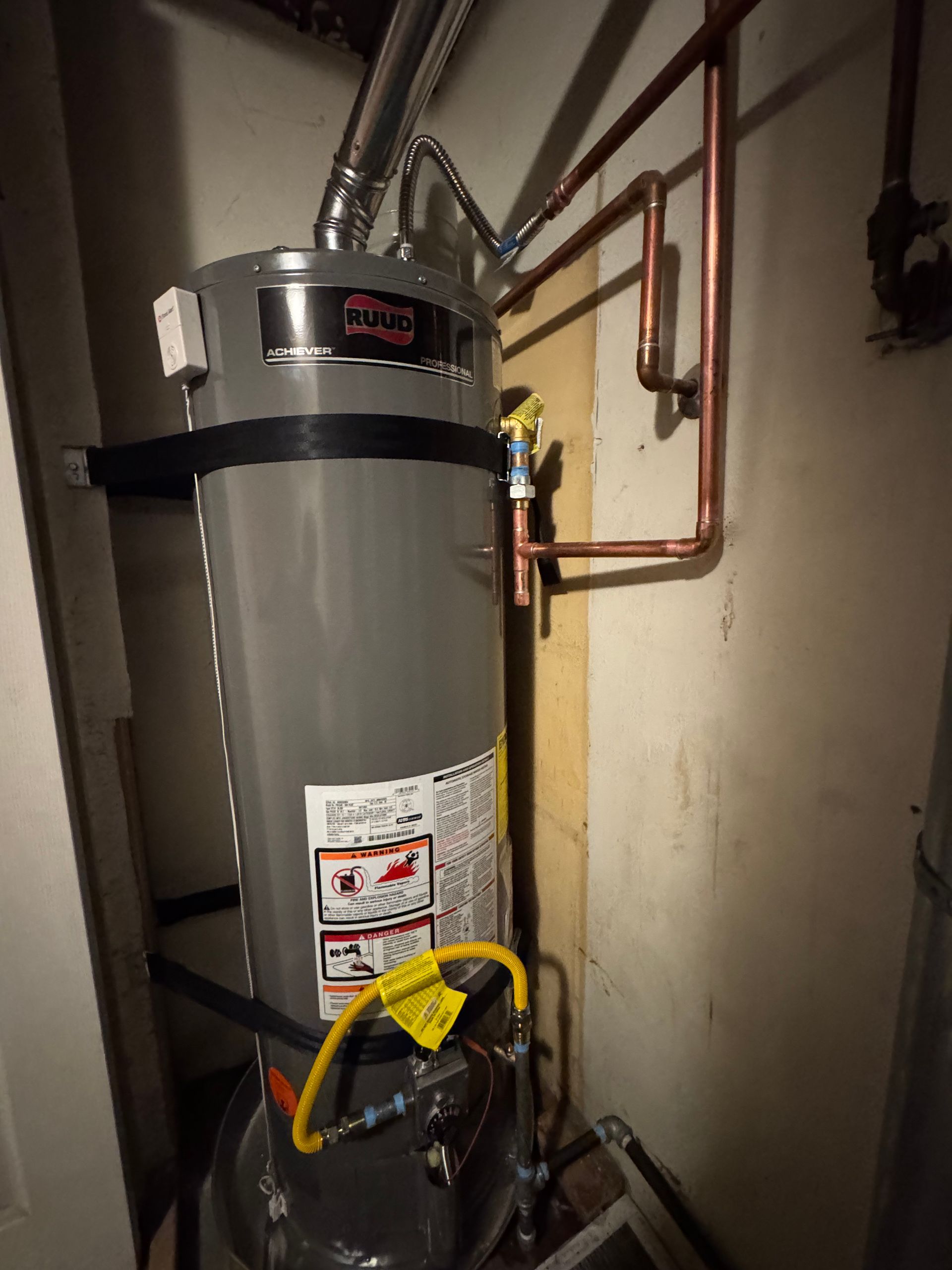 A gas water heater is sitting in a corner of a room next to copper pipes.