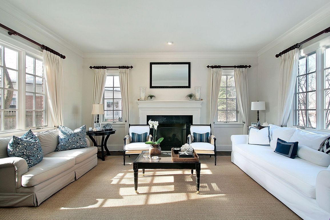 Bright living room with white sofas, coffee table, fireplace, and large windows with patterned curtains Bright living room with white sofas, coffee table, fireplace, and large windows with patterned curtains