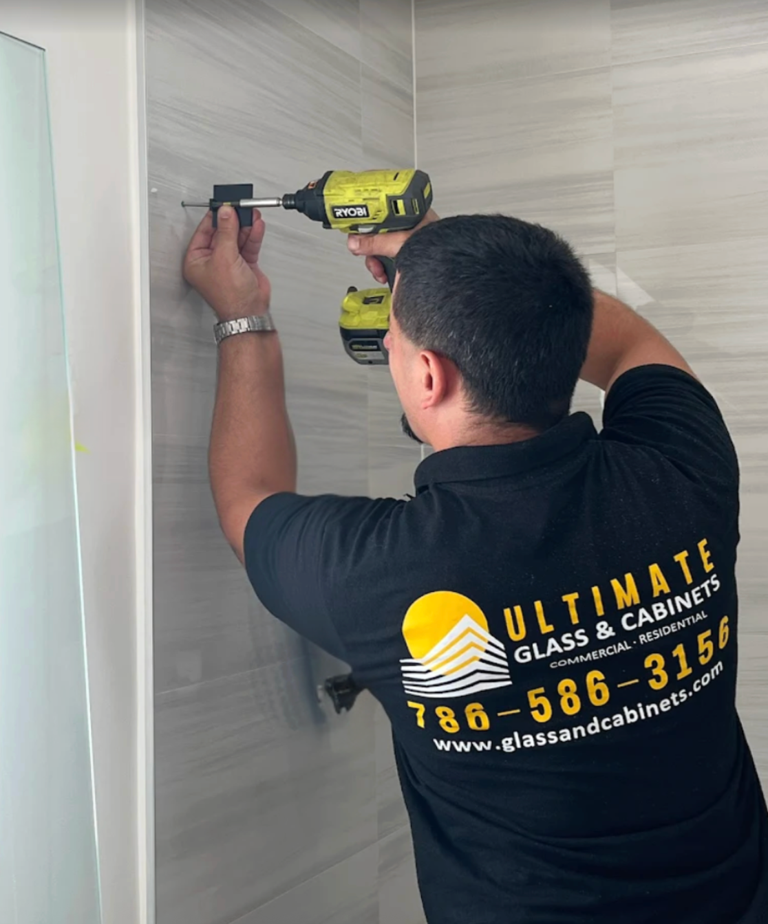 Person uses a drill to install hardware on a tiled wall. The worker wears a black shirt with a company logo.