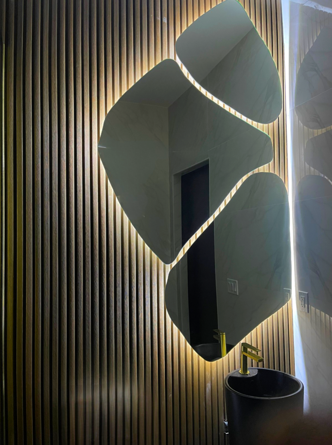 Mirror with internal lighting, mounted on ribbed wall above a dark sink and gold faucet.
