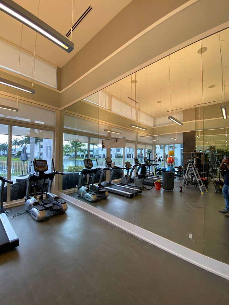 Gym with treadmills, weights, and mirrors. Natural light floods the space. A person works on equipment.