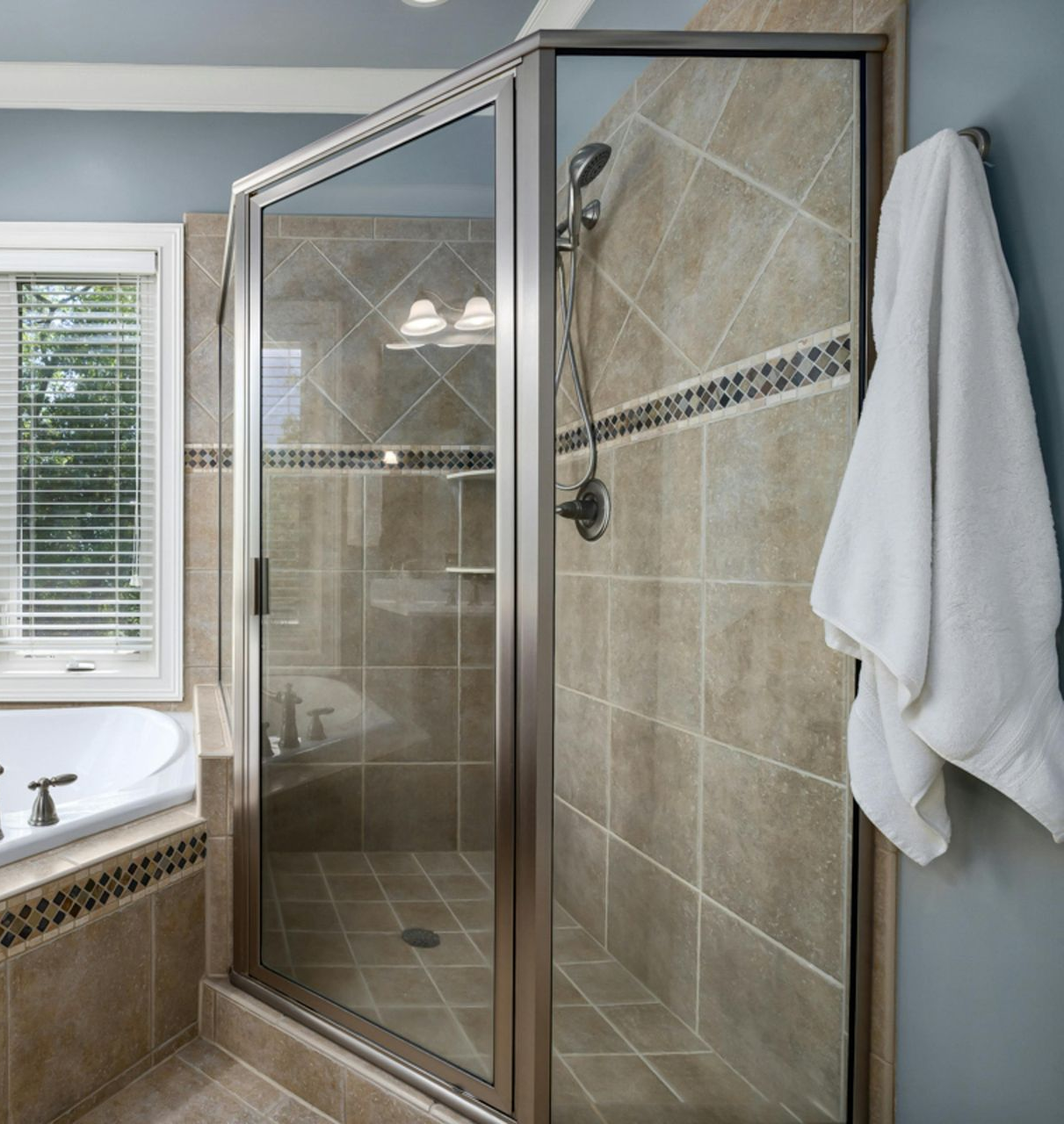 A glass-doored shower in a bathroom with beige tile, a white towel, and a window.