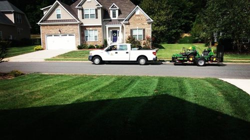 A white truck is towing a green tractor in front of a house.