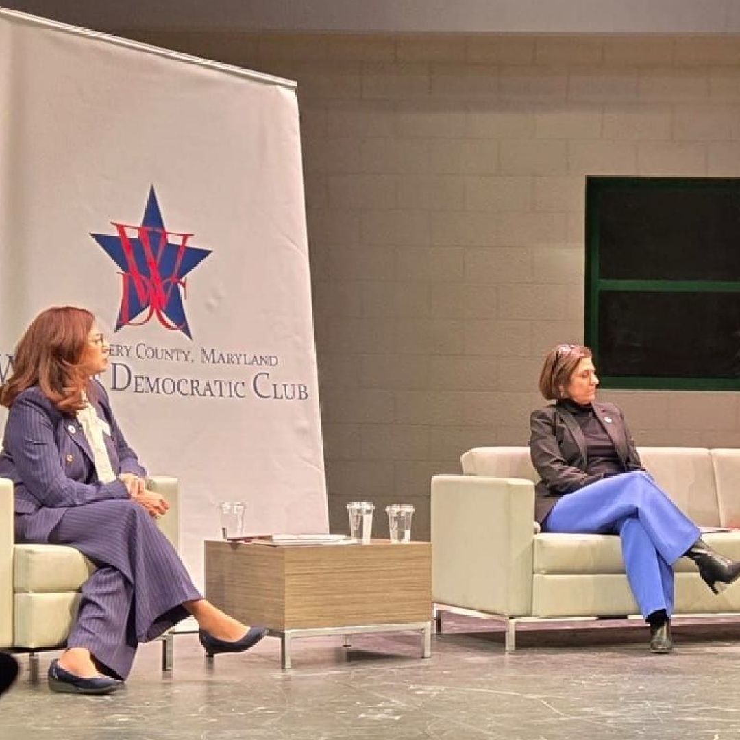 Two women are sitting on couches in front of a democratic club banner.
