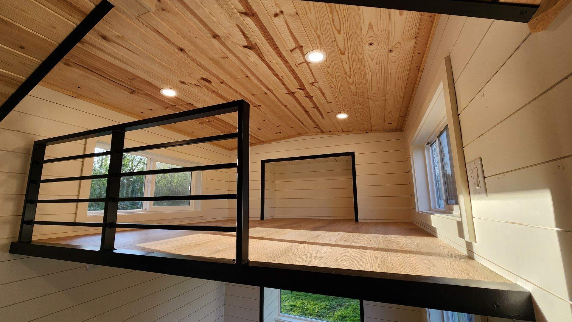 Interior of a small home, featuring a loft with a black railing, wooden walls and ceiling.