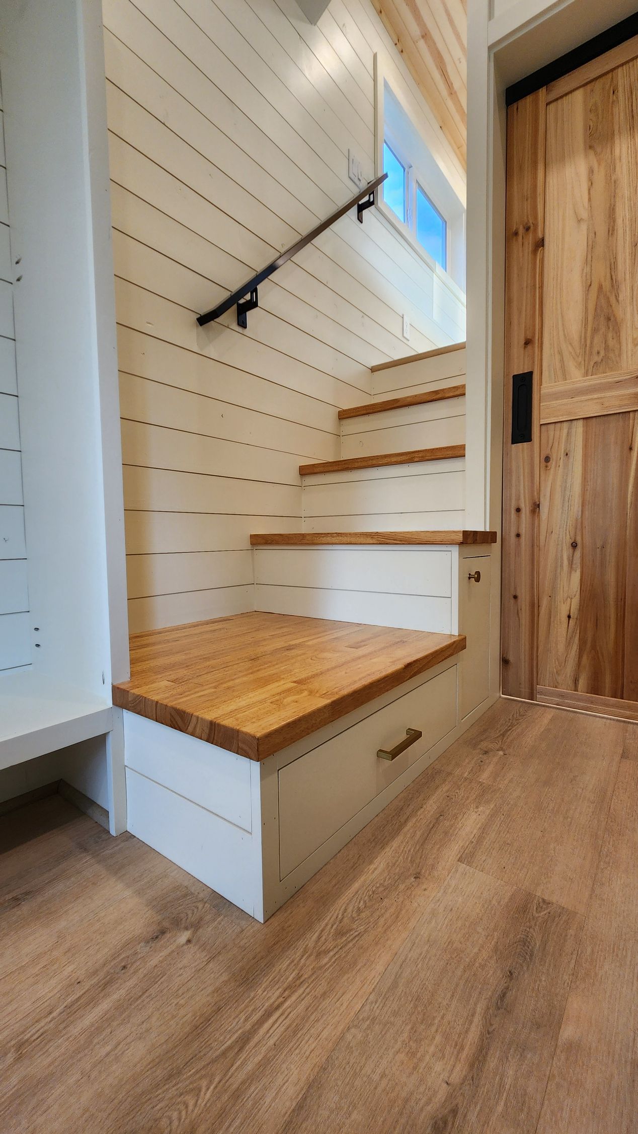 Staircase with built-in drawers in tiny home. Wood and white paneling, wooden door and handrail.