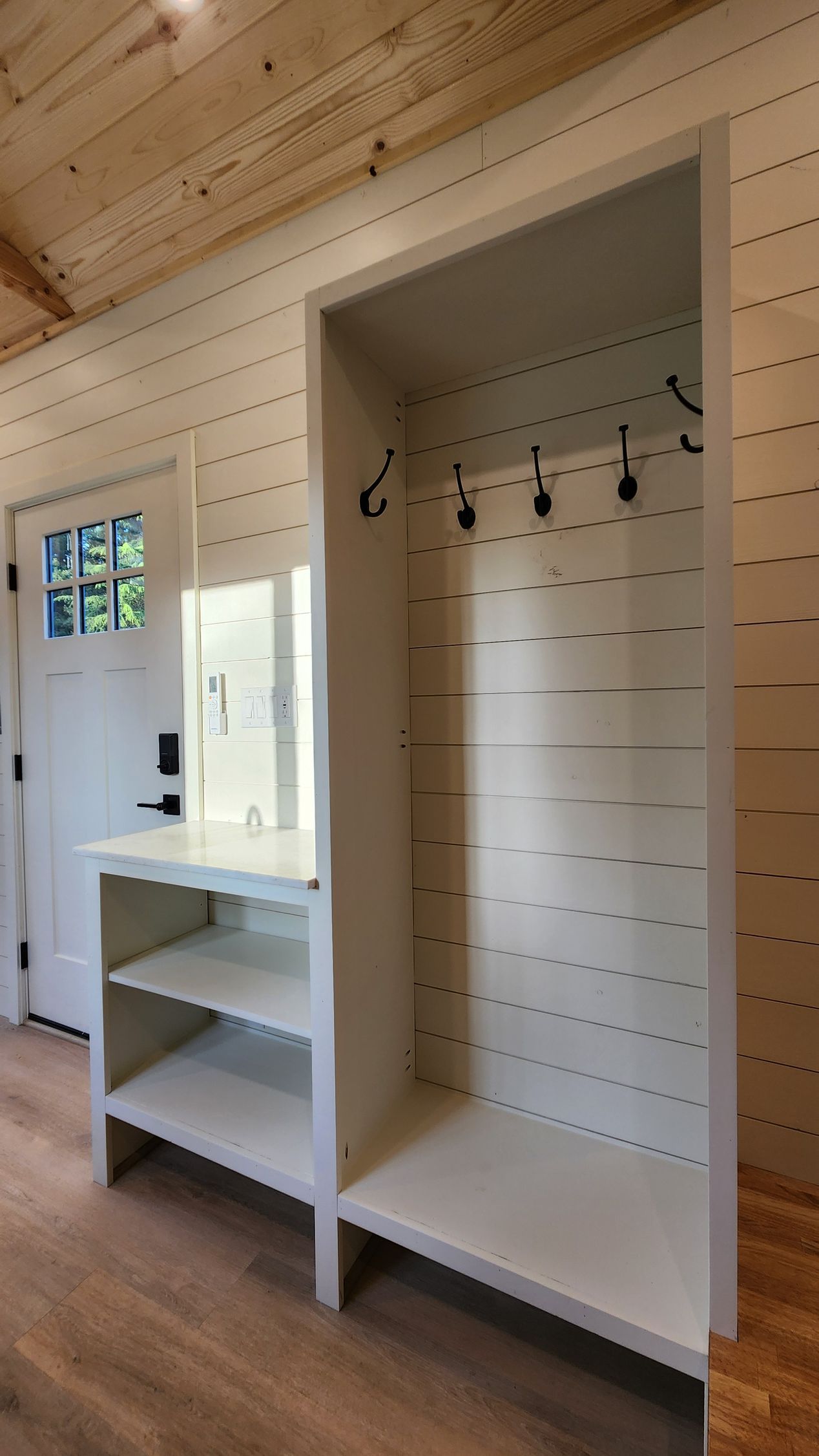 White built-in entryway storage with shelves, hooks, and a mirror, next to a door in a light-colored room.