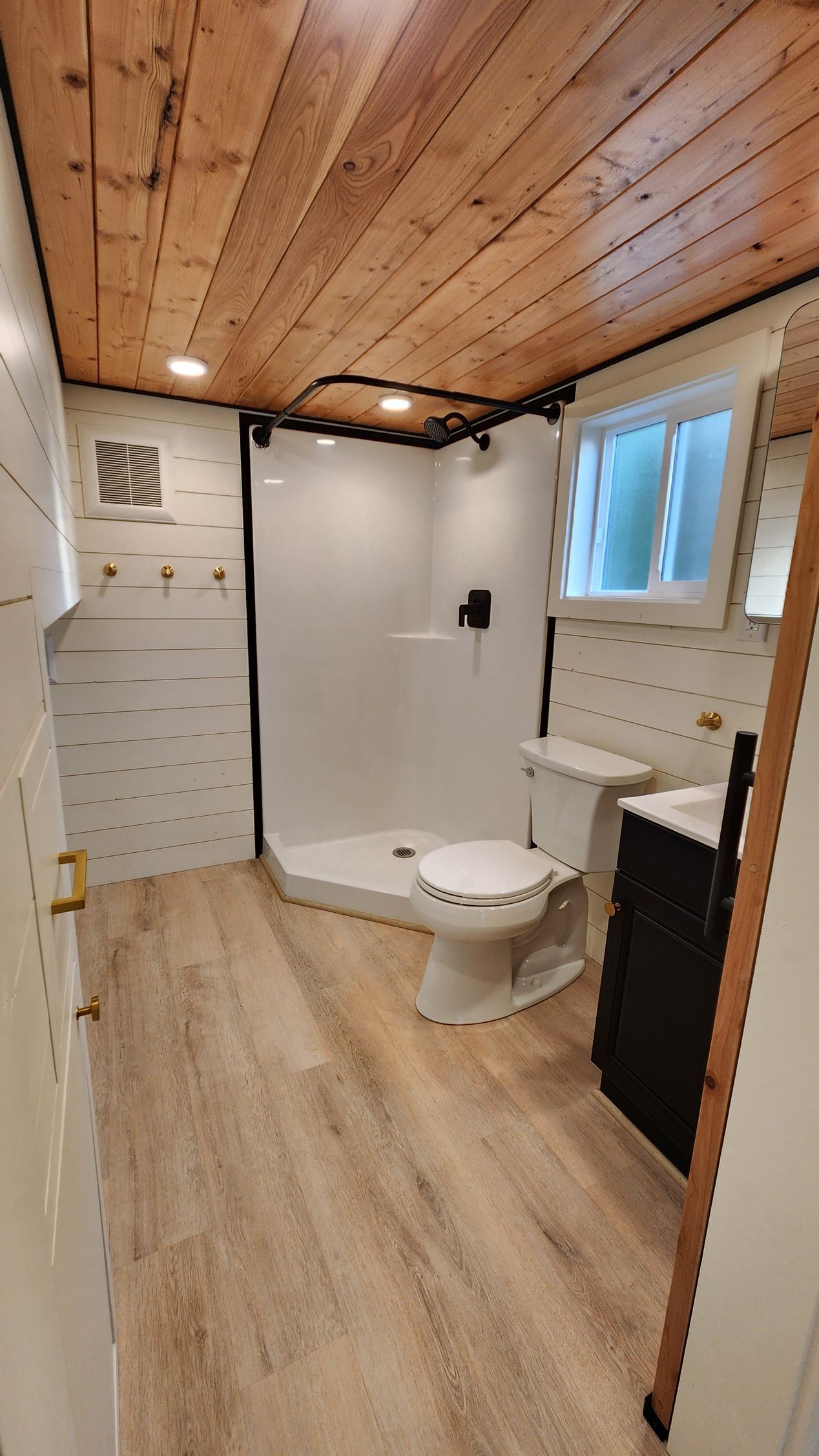 Bathroom with light wood floor and ceiling, white shower, toilet, and walls with black accents. Window, sink, cabinet.