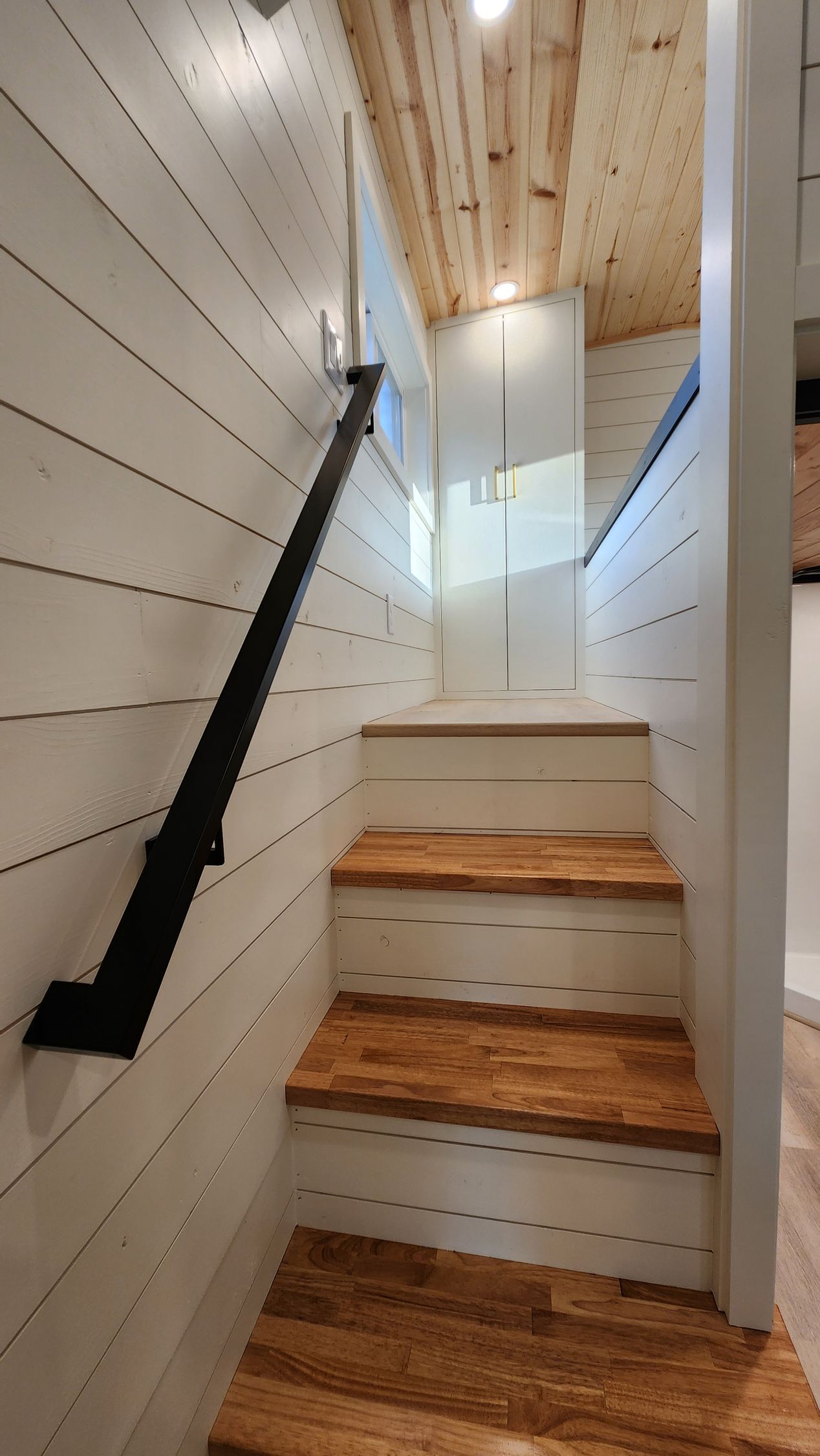 Staircase with wood treads and a black handrail, leading up to a white door and loft in a small space.