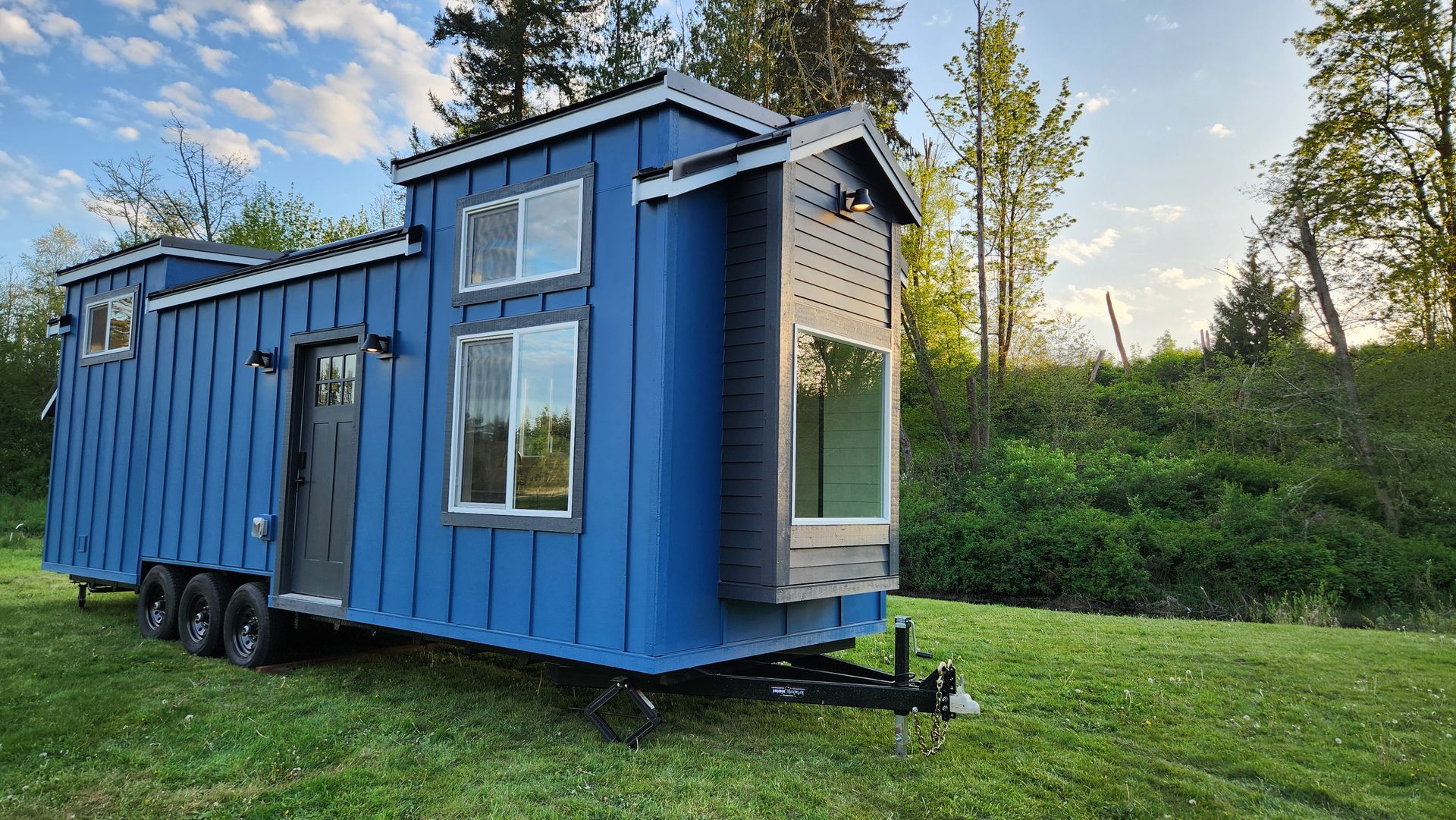 Blue tiny house on wheels in a grassy field with a forest background.