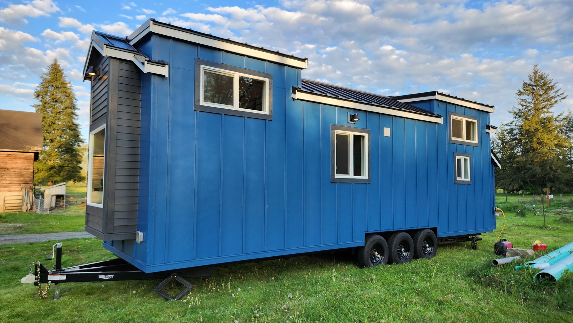 Blue tiny house on a trailer in a grassy field, with black accents and multiple windows.