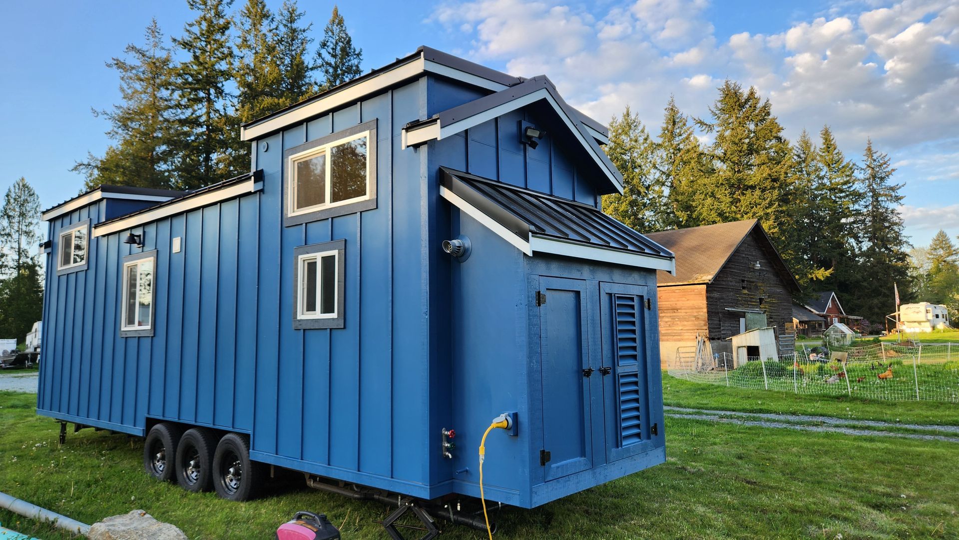 Blue tiny house on wheels with black roof, parked in a grassy yard.