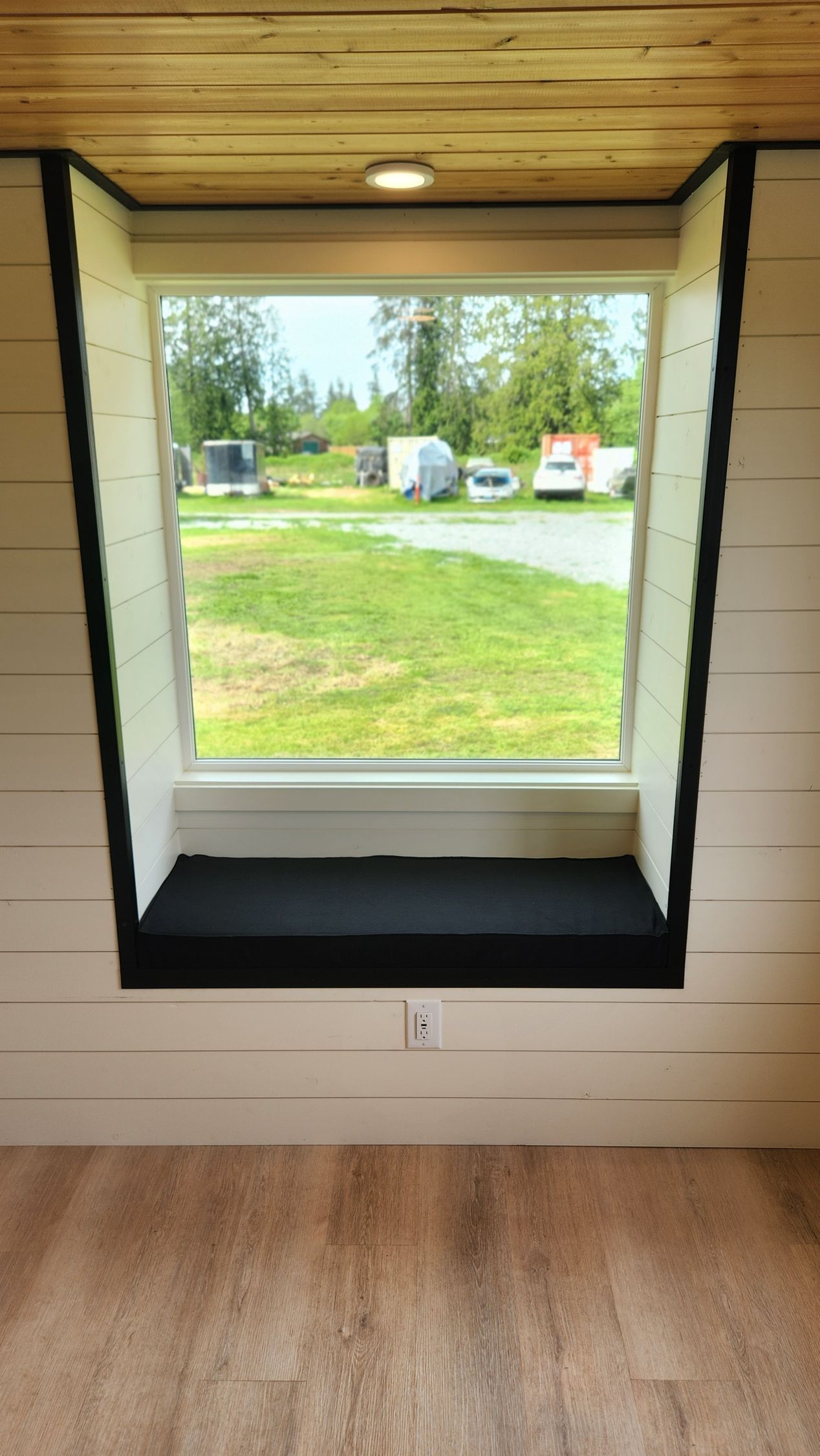Window seat inside a tiny home, looking out over a grassy field. Black trim and white walls, wooden floor.