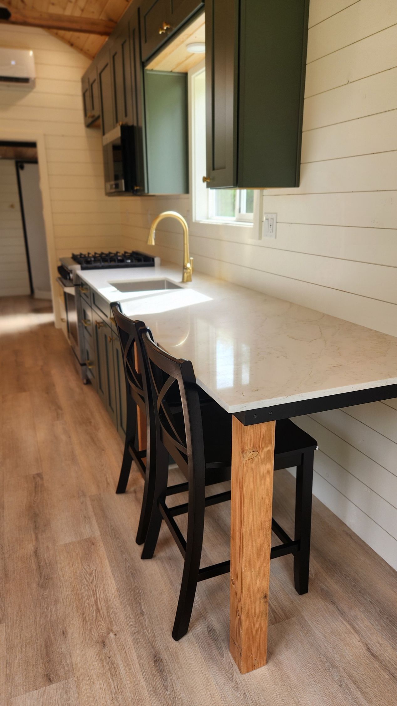 Kitchen with a long countertop, black cabinets, gold faucet, and wood accents. Two black bar stools are at the counter.