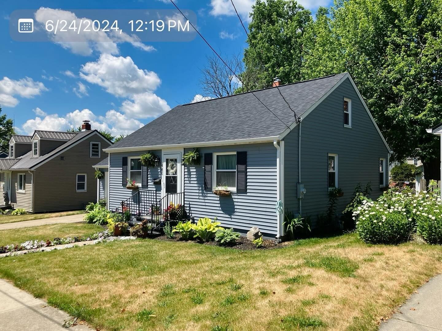 Small blue-gray house with white trim, front porch, and a dry lawn under a partly cloudy sky