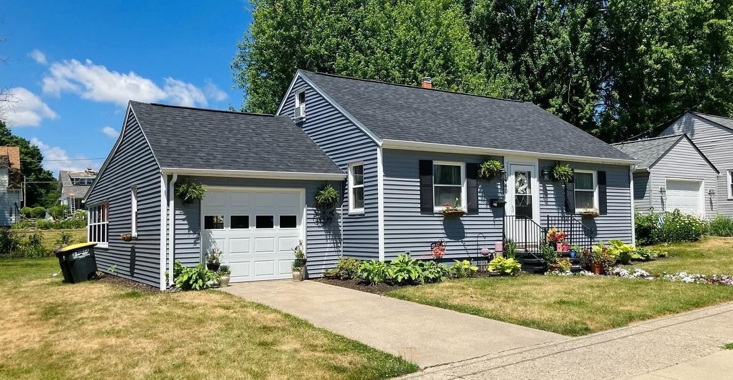 Gray house with attached garage, front porch, and flower beds on a sunny suburban street