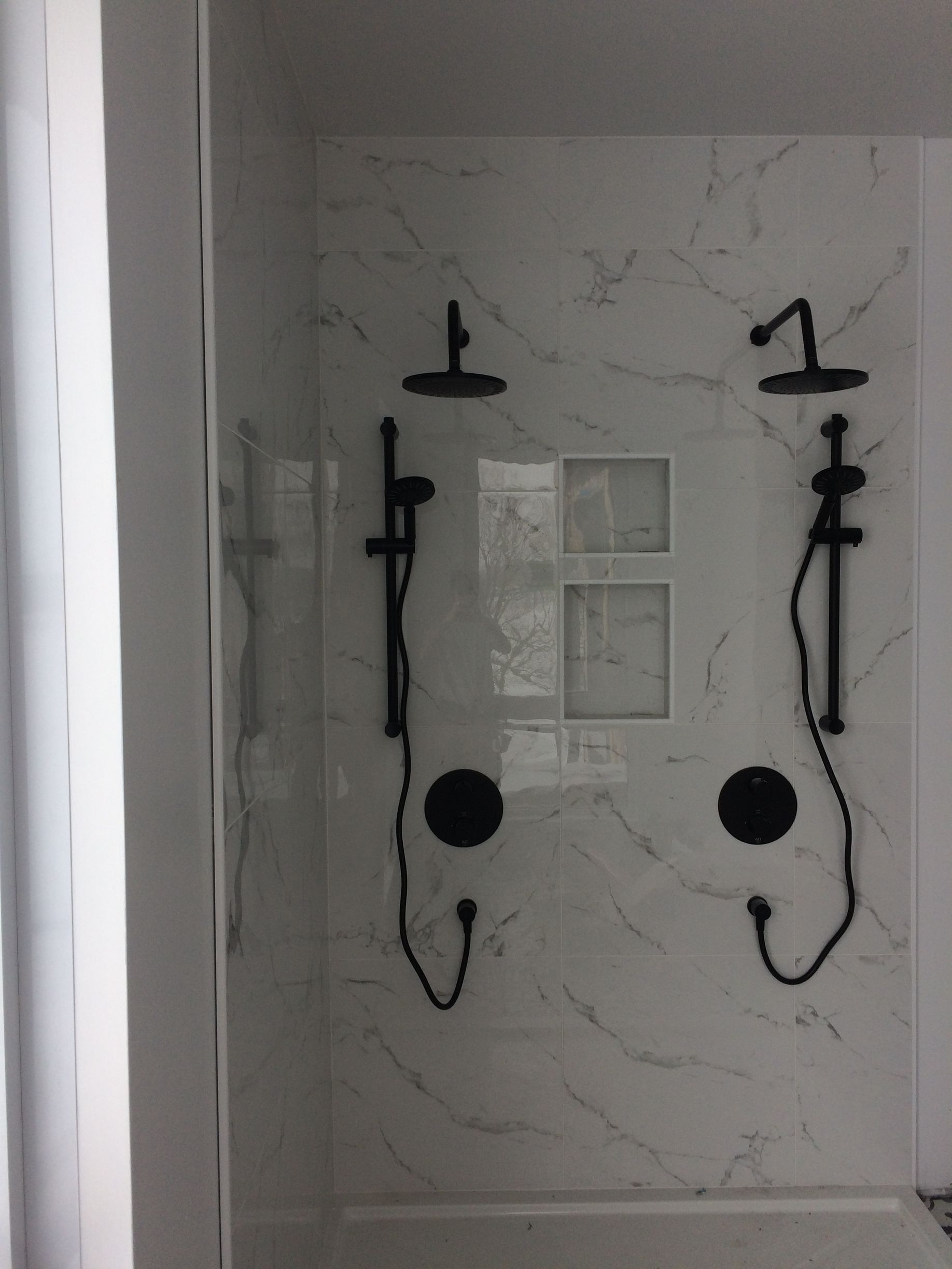 Robinetterie de douche noire sur un mur carrelé de marbre blanc. Deux pommeaux de douche de tête et douchettes à main.