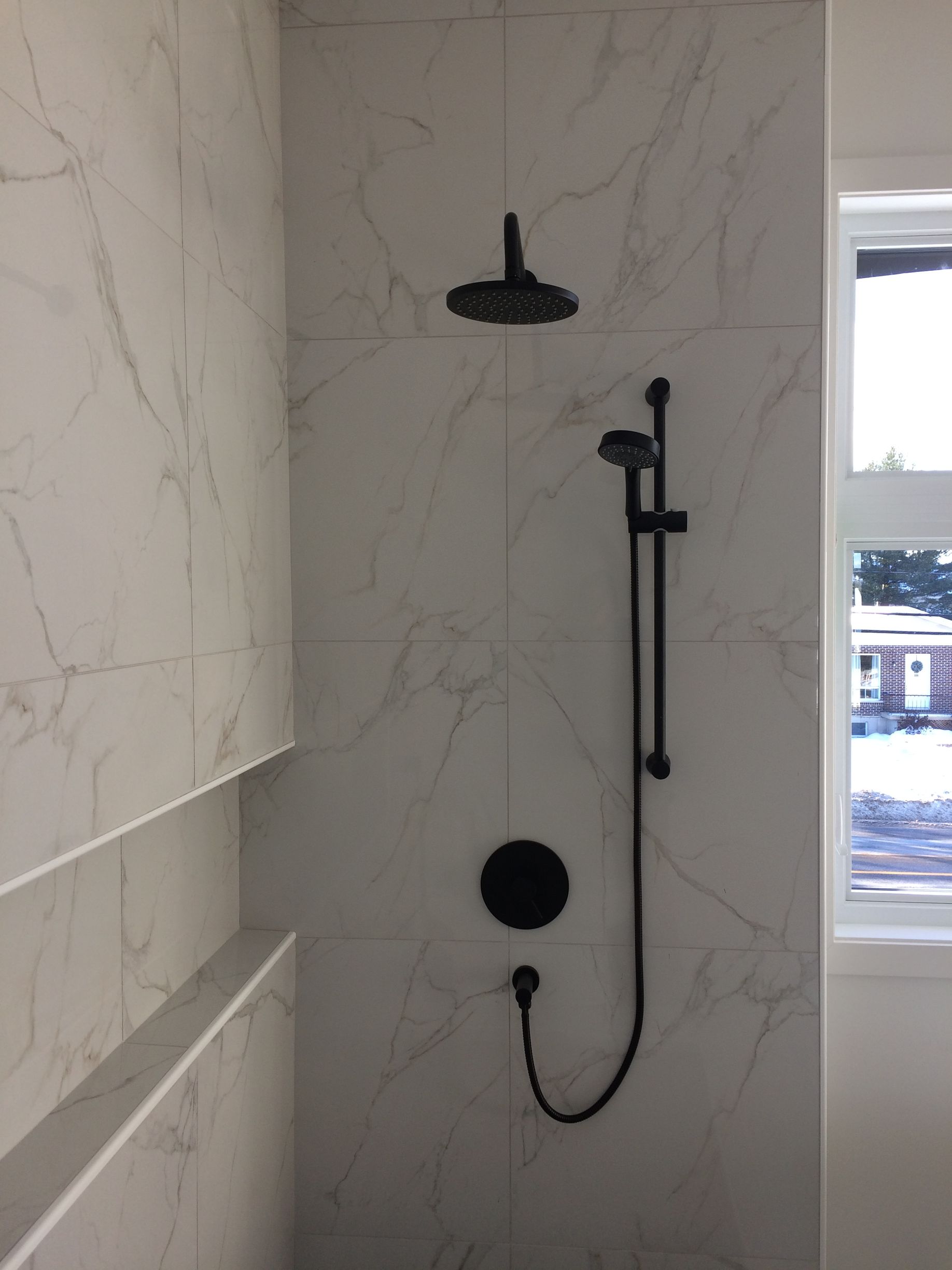 Douche avec robinetterie noire et carrelage imitation marbre. Comprend une pomme de douche effet pluie et une douchette.