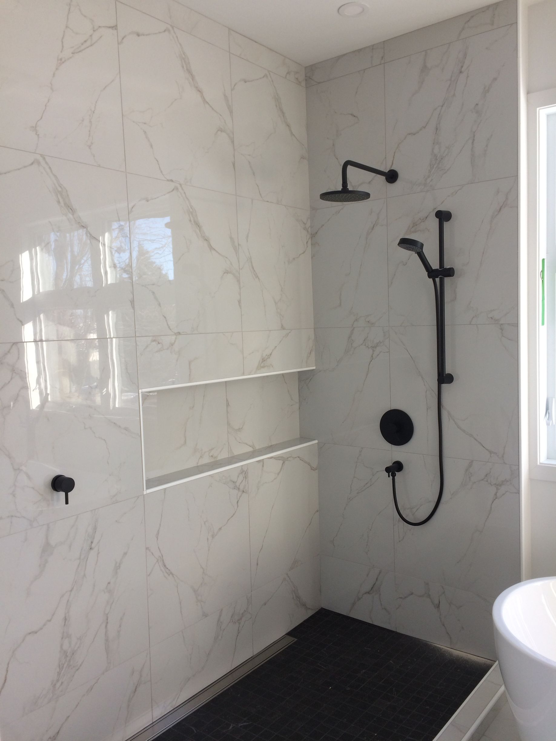 Douche avec carrelage imitation marbre blanc, robinetterie noire, étagères encastrées et carrelage noir au sol.